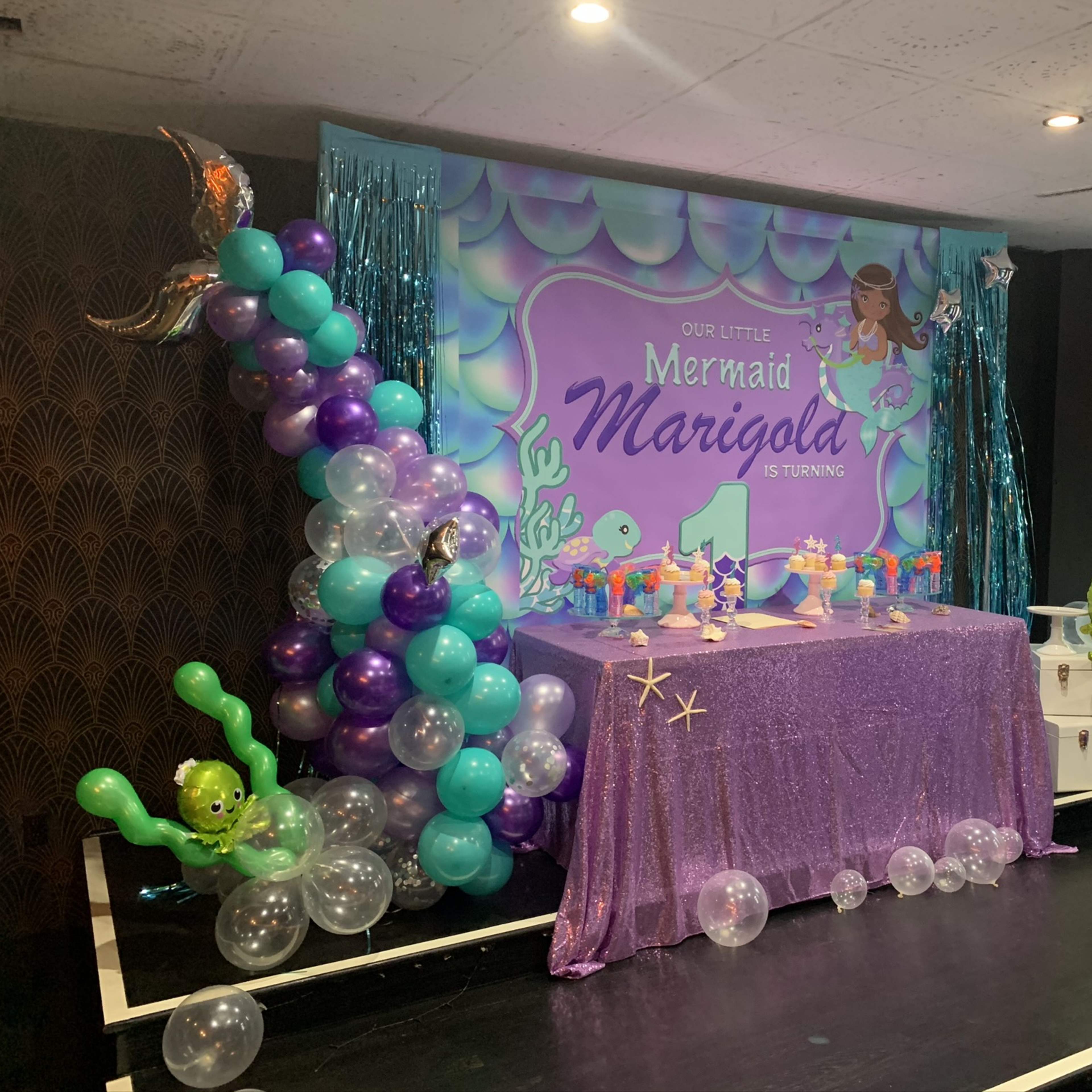 A colorful birthday party setup with a mermaid theme, featuring balloons, a decorated cake table, and a backdrop displaying "Our Little Mermaid Marigolda is Turning 1."