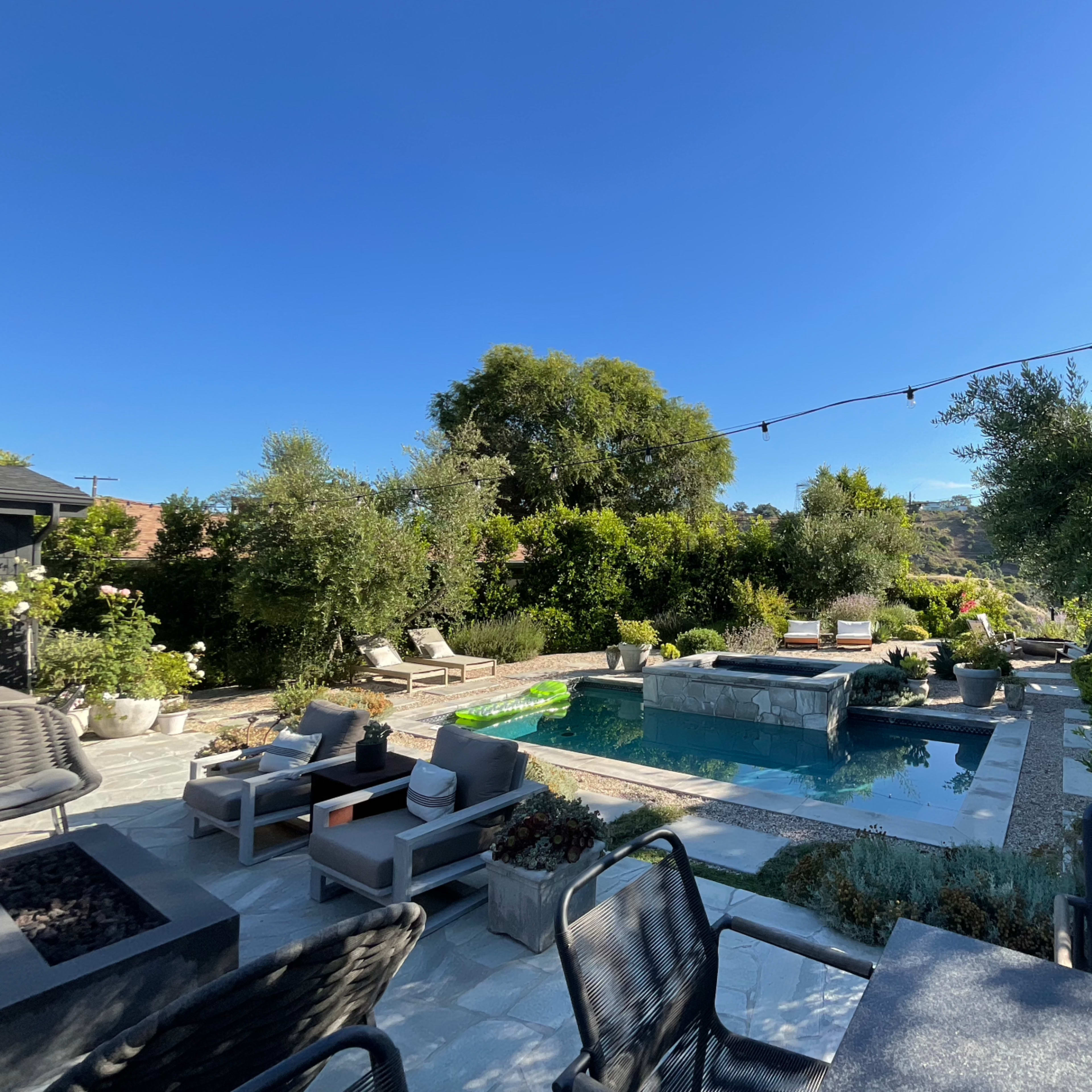 A backyard featuring a swimming pool, lounge chairs, and landscaped greenery under a clear blue sky.