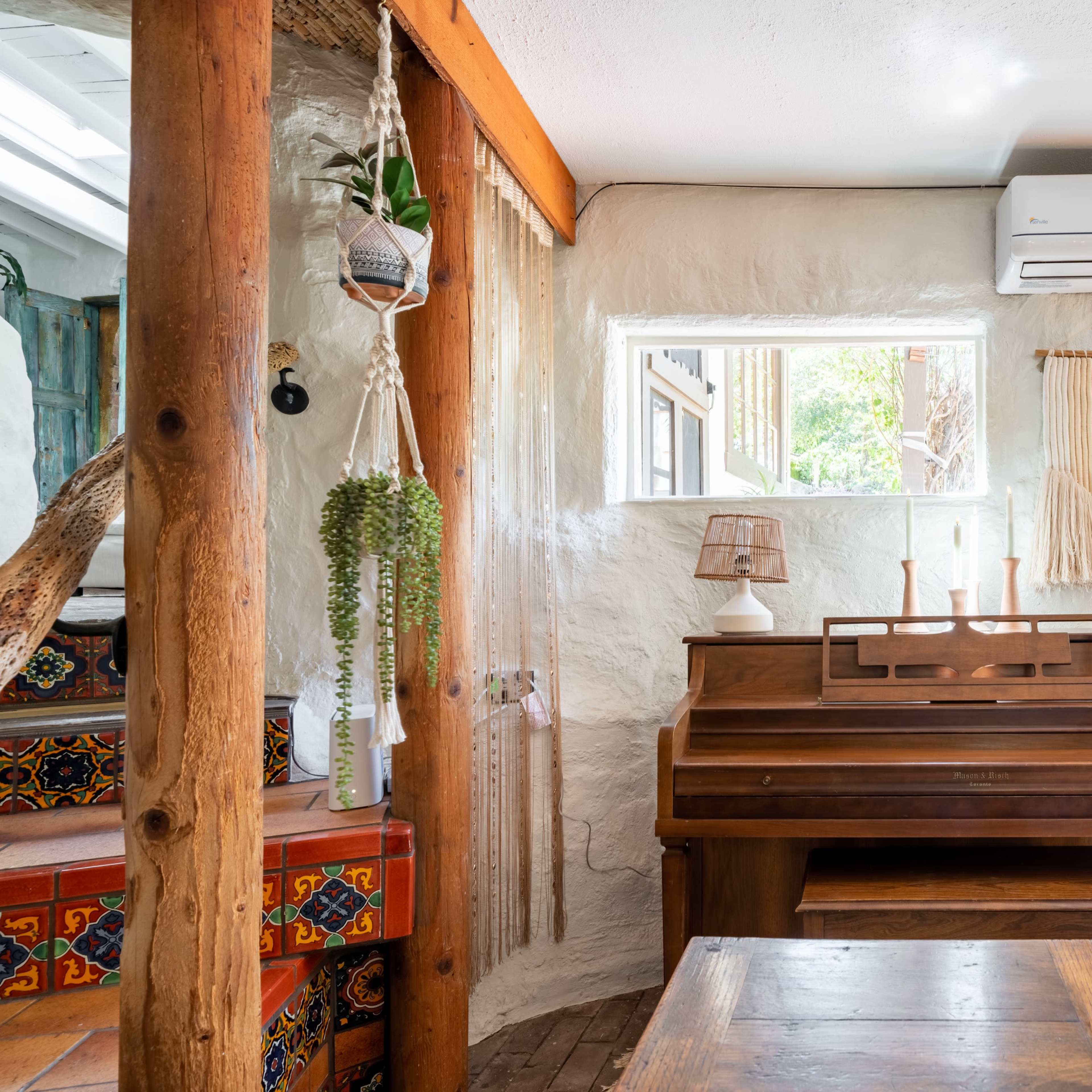 A wooden piano sits in a cozy room with natural light streaming through the windows, surrounded by decorative elements and wooden beams.