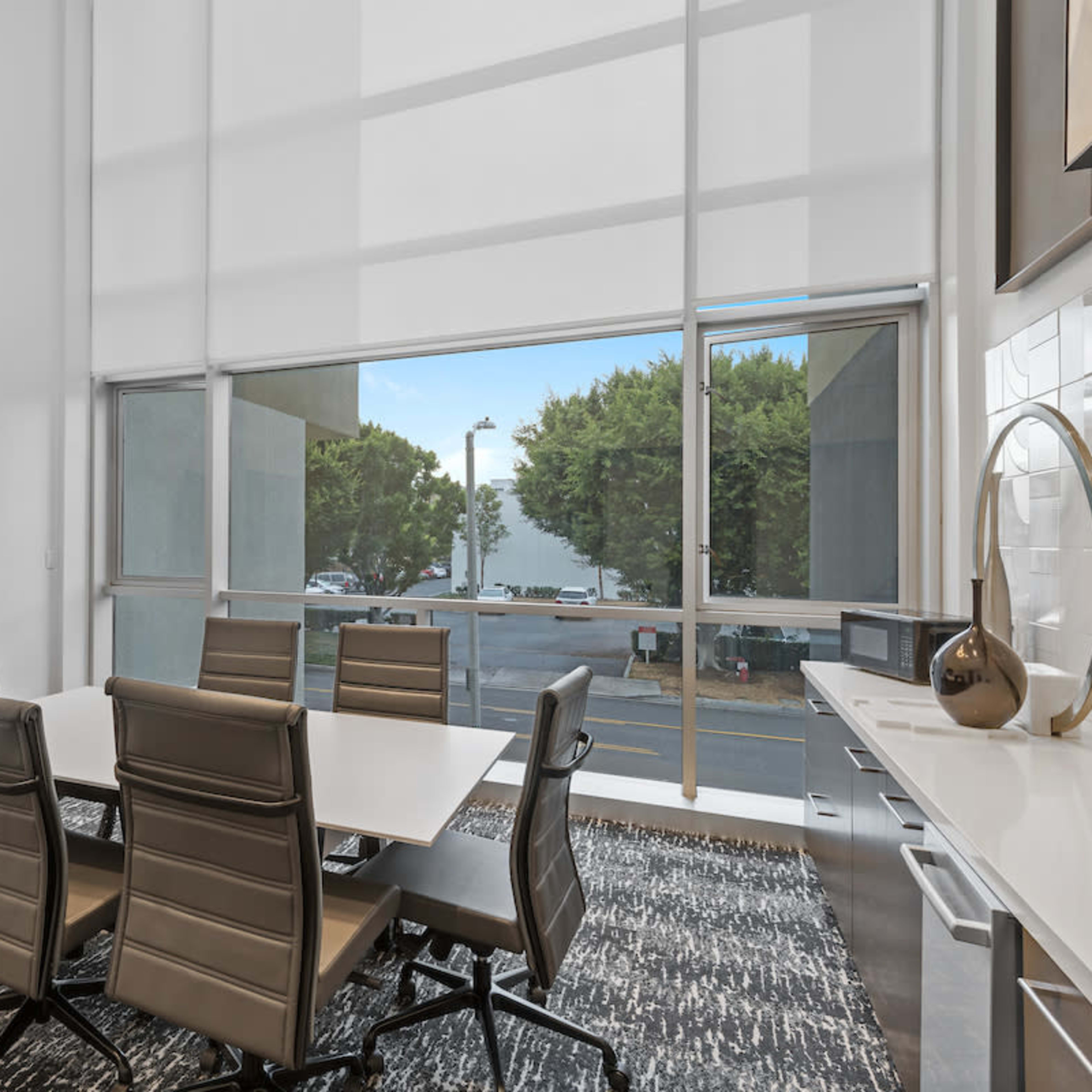 A modern conference room features a large window, a rectangular table surrounded by six leather chairs, and a wall-mounted television.