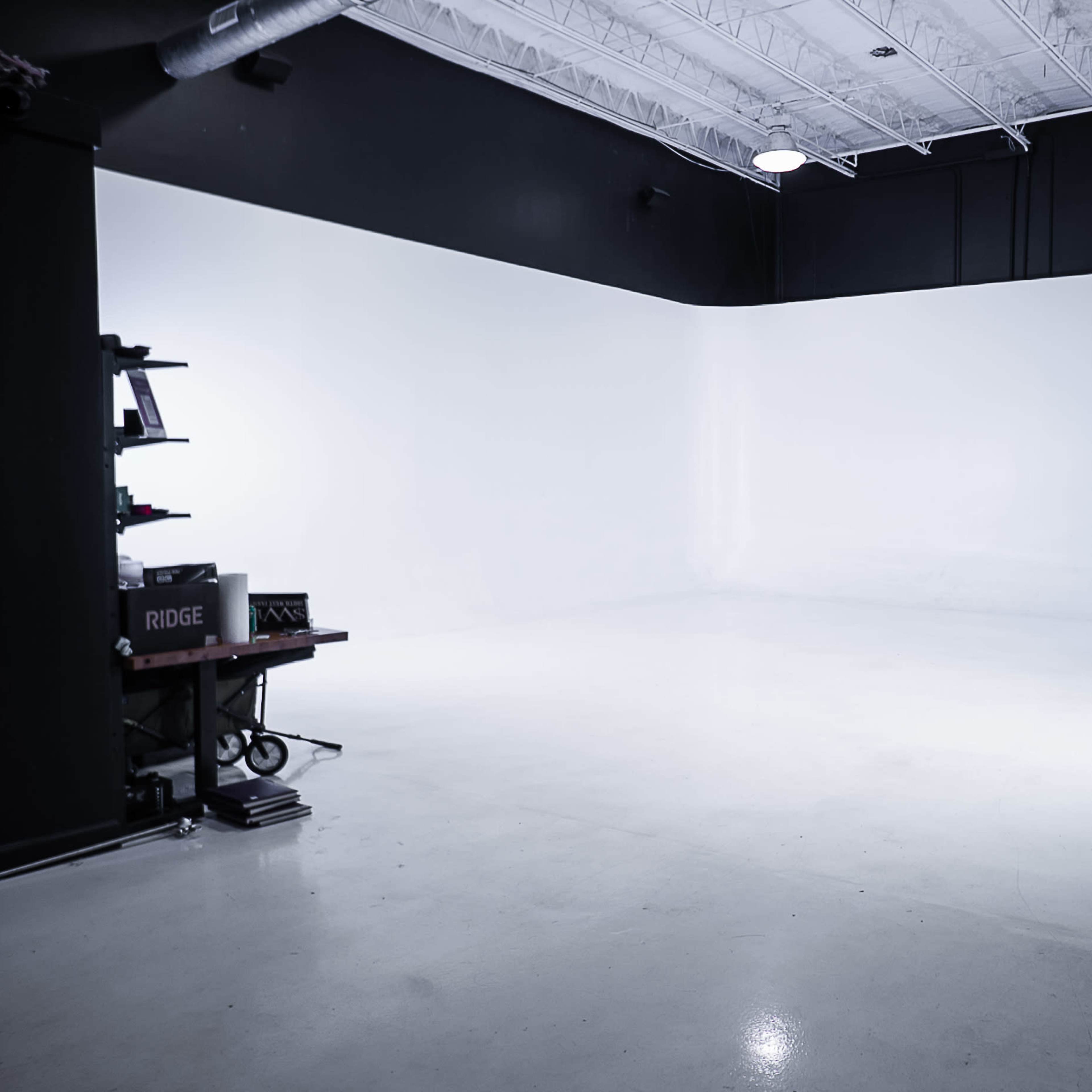 A spacious photography studio with a plain white backdrop, concrete flooring, and minimal equipment arranged along the side.