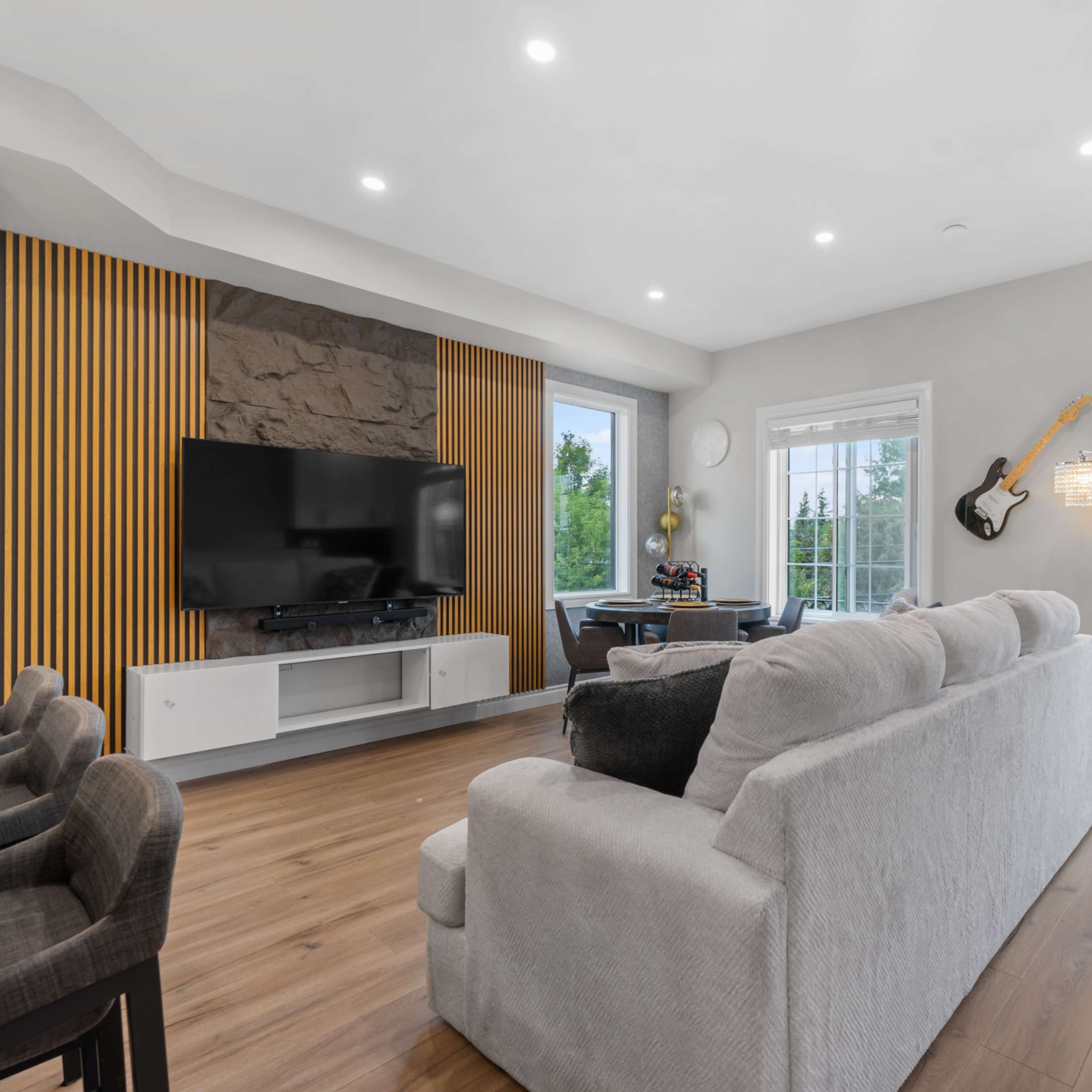 The living room features a television mounted on a stone wall, with large windows overlooking greenery and a guitar hanging on the wall.