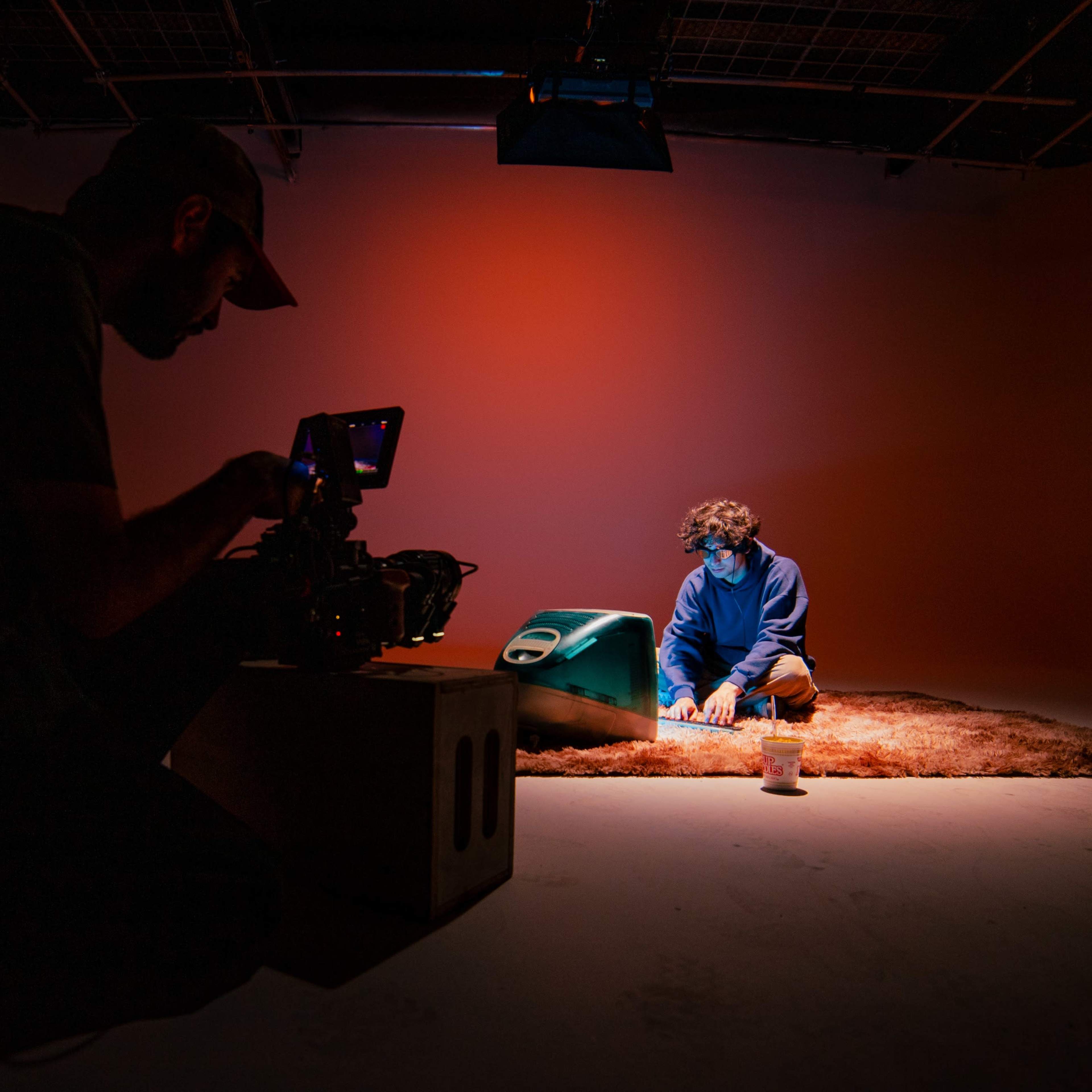 A person sits on a rug in a dimly lit studio, while another person operates a camera, capturing the scene.