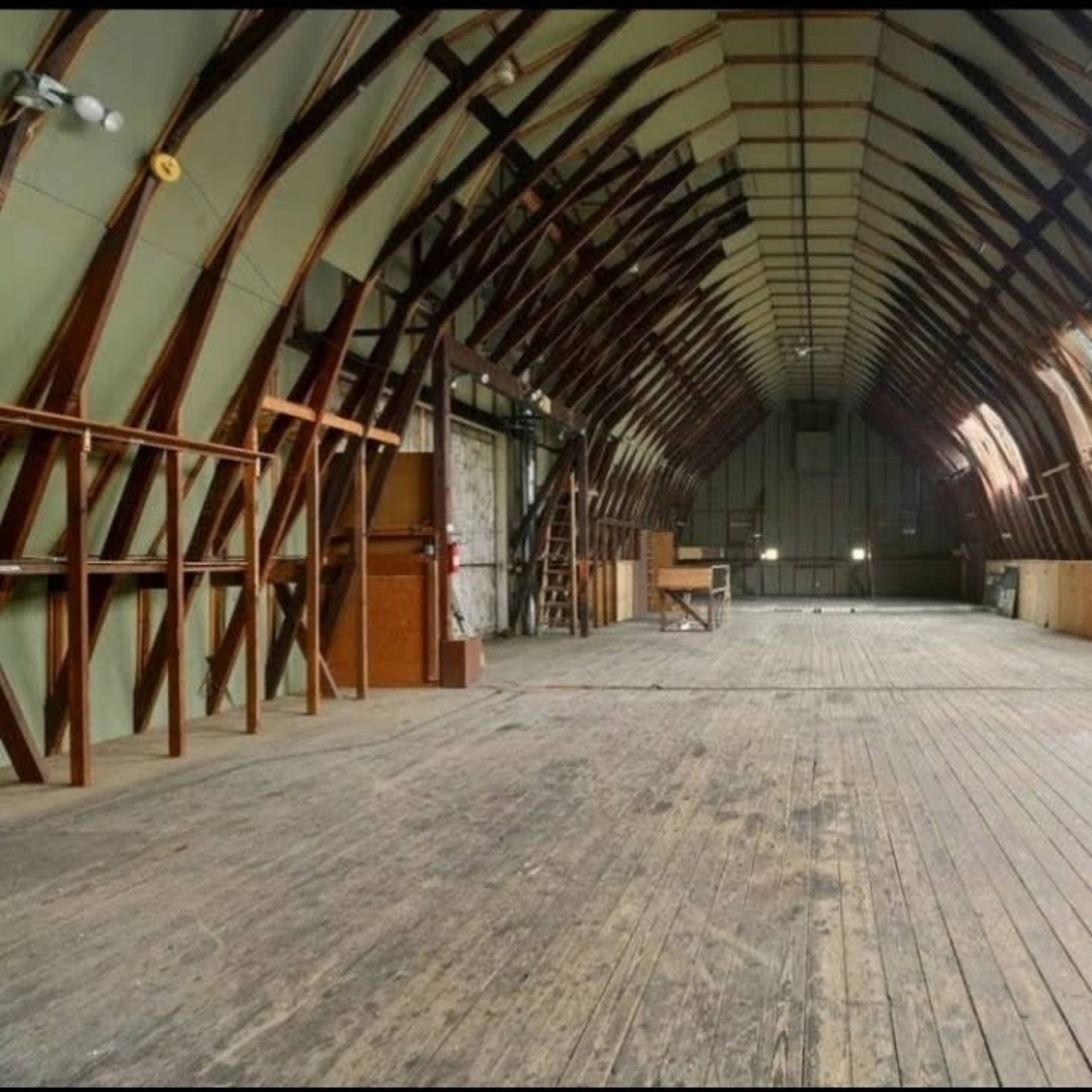 A spacious, unfinished interior of a building with high arched wooden beams and a wooden floor.