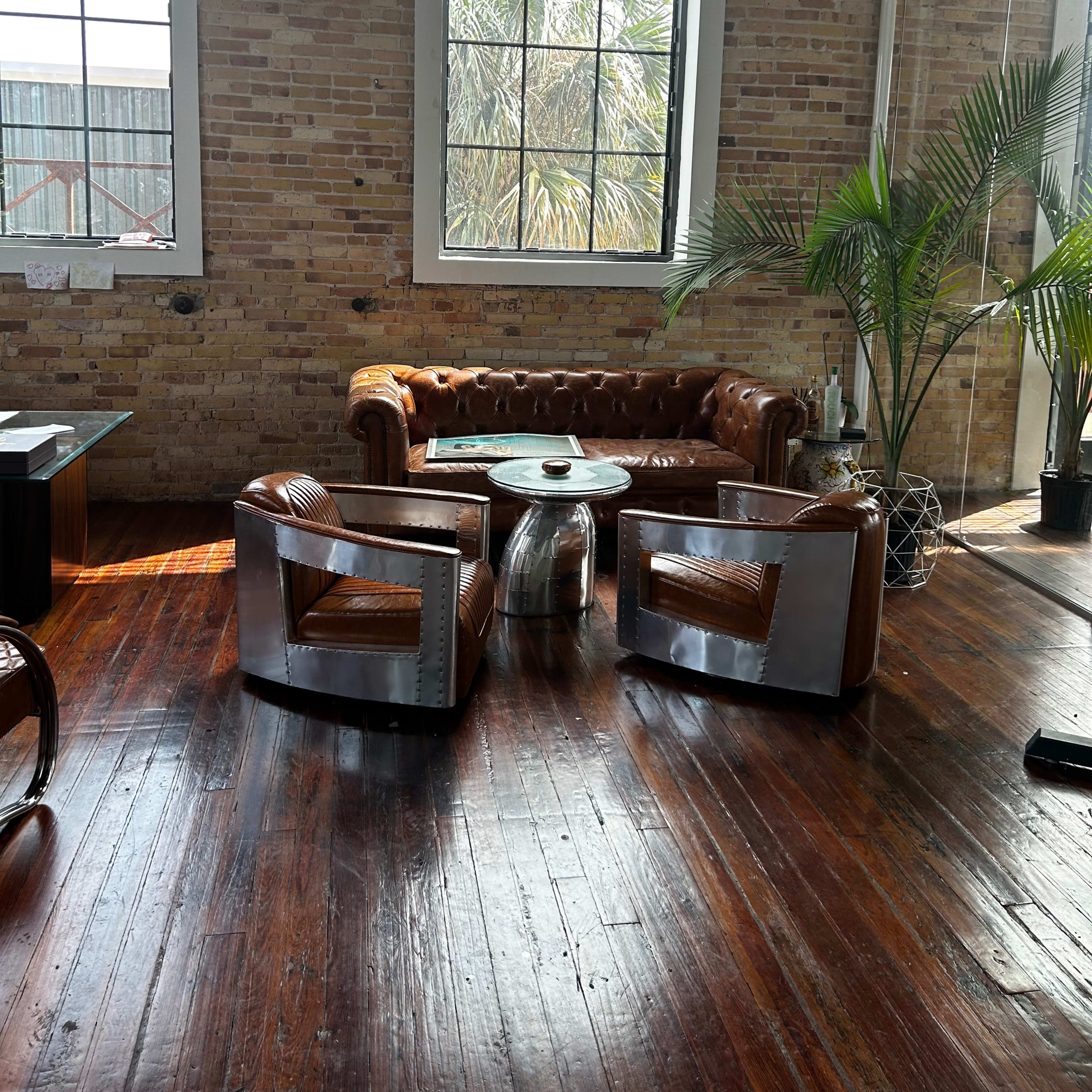 The scene features a leather chesterfield sofa and two contemporary chairs surrounded by large windows and a palm plant in a room with exposed brick walls and polished wooden floors.