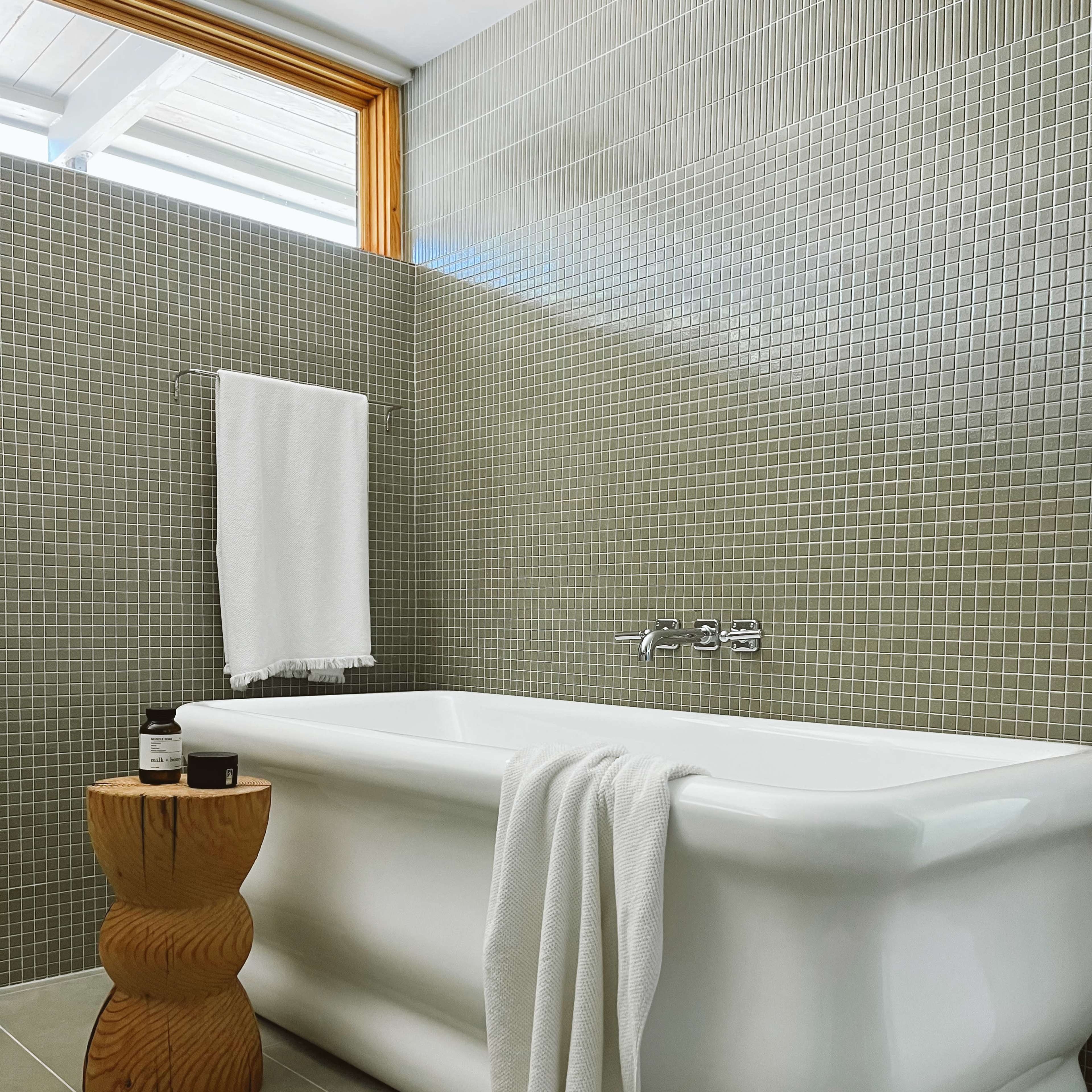 A modern bathroom with a white freestanding bathtub, a wooden stool, and green mosaic tiled walls.