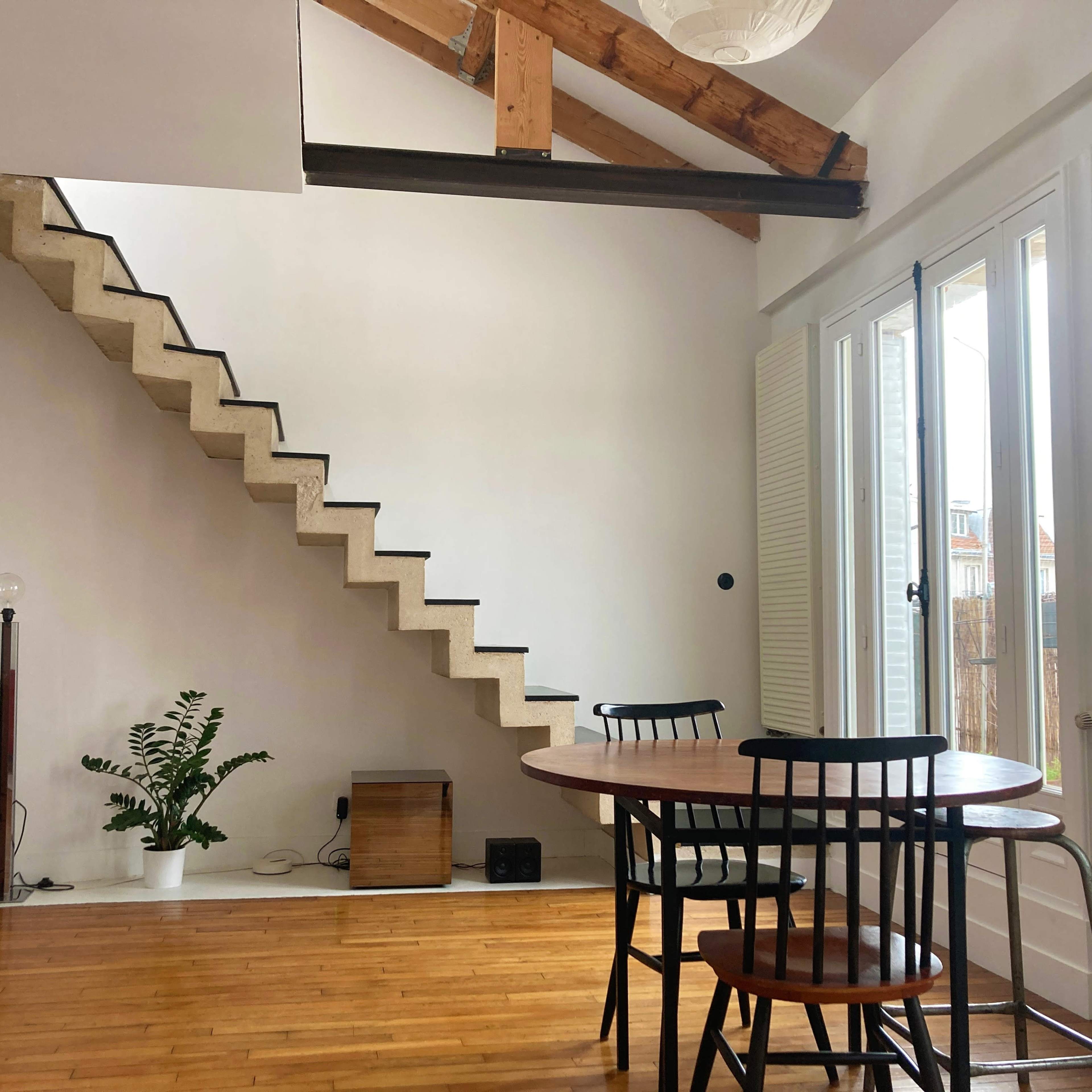 A modern interior features a round dining table with black chairs, a staircase leading to an upper level, and large windows providing natural light.