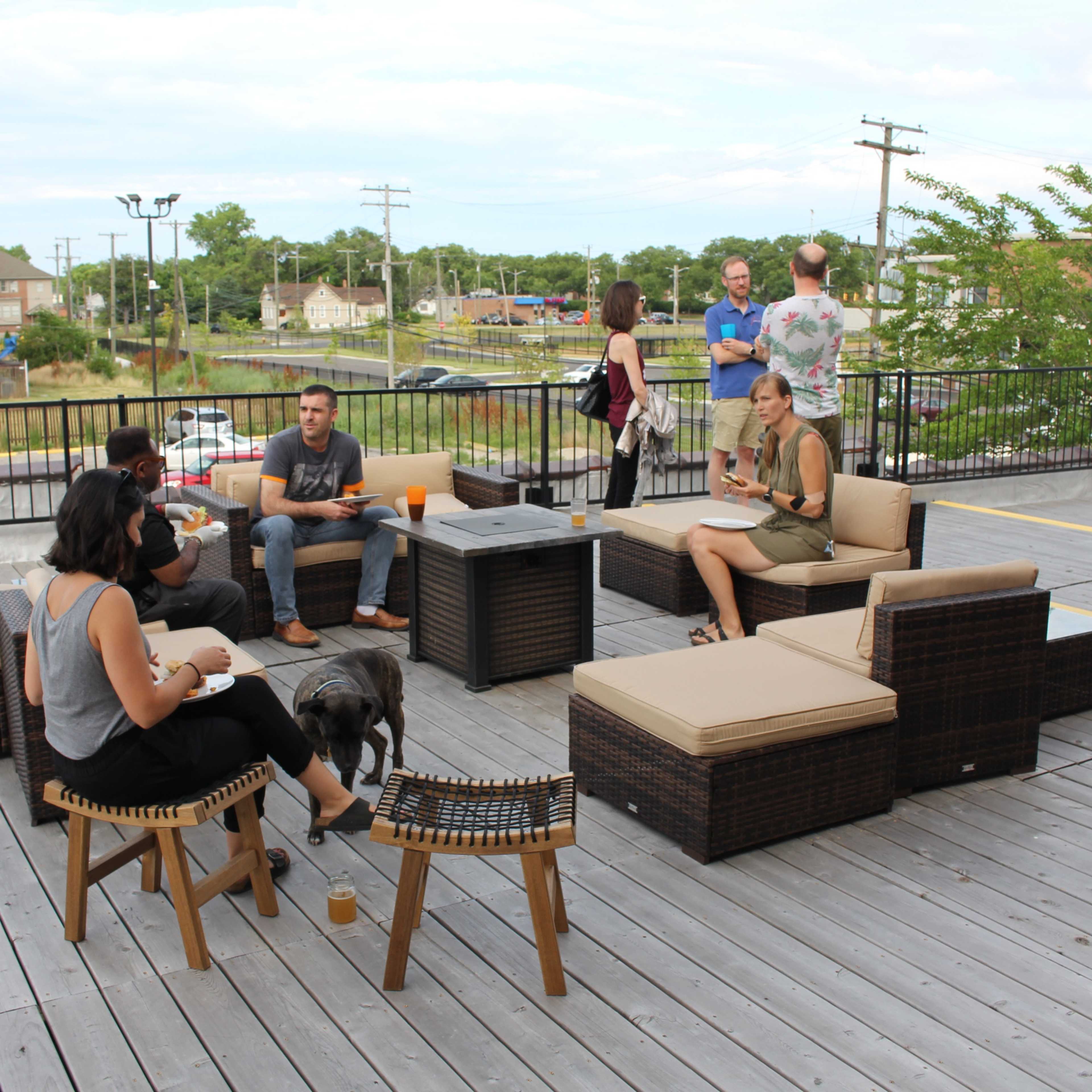 A group of people relaxes on a patio furnished with wicker chairs and a central table, with some enjoying food and drinks.