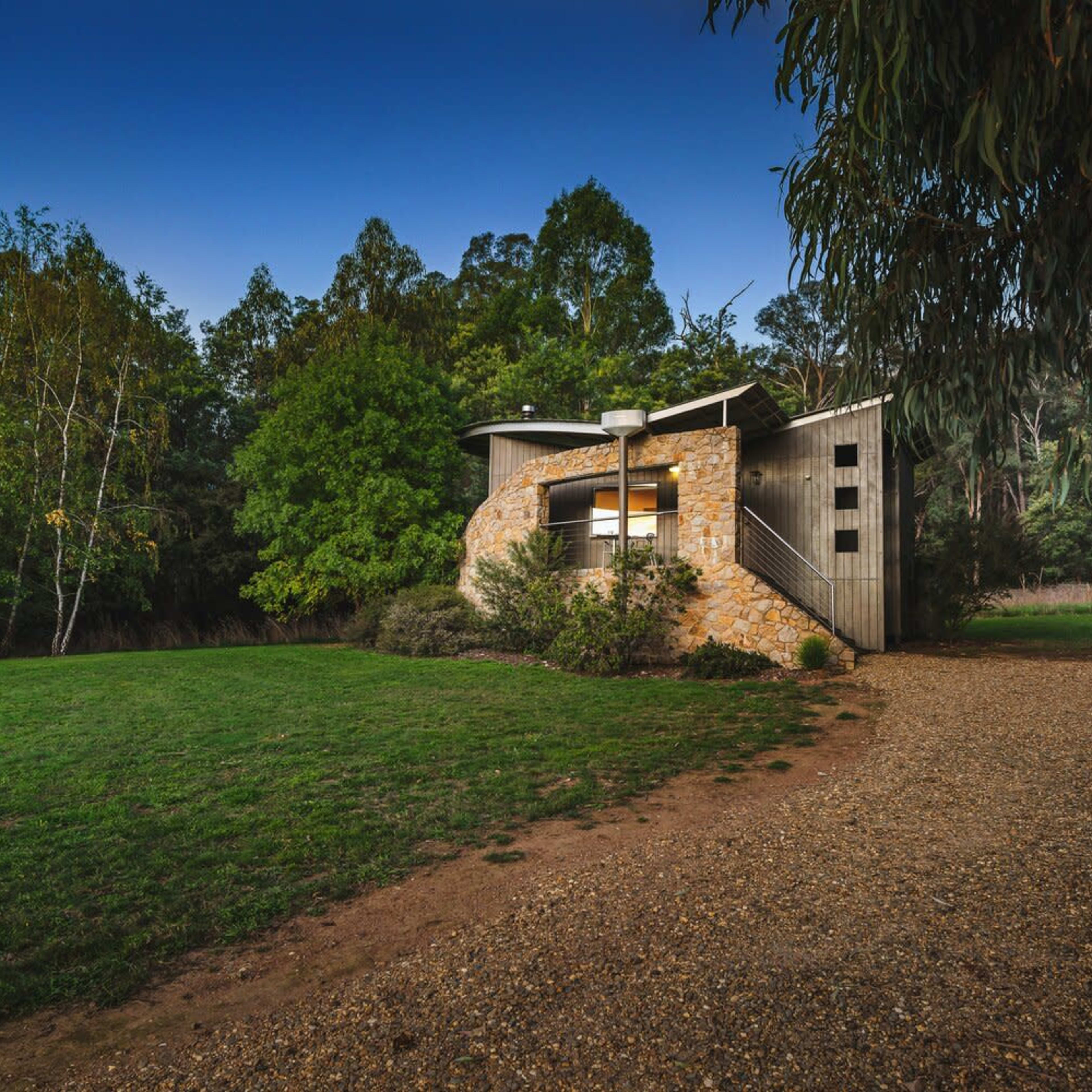 A modern house with a stone facade is situated on a gravel path surrounded by trees and greenery.