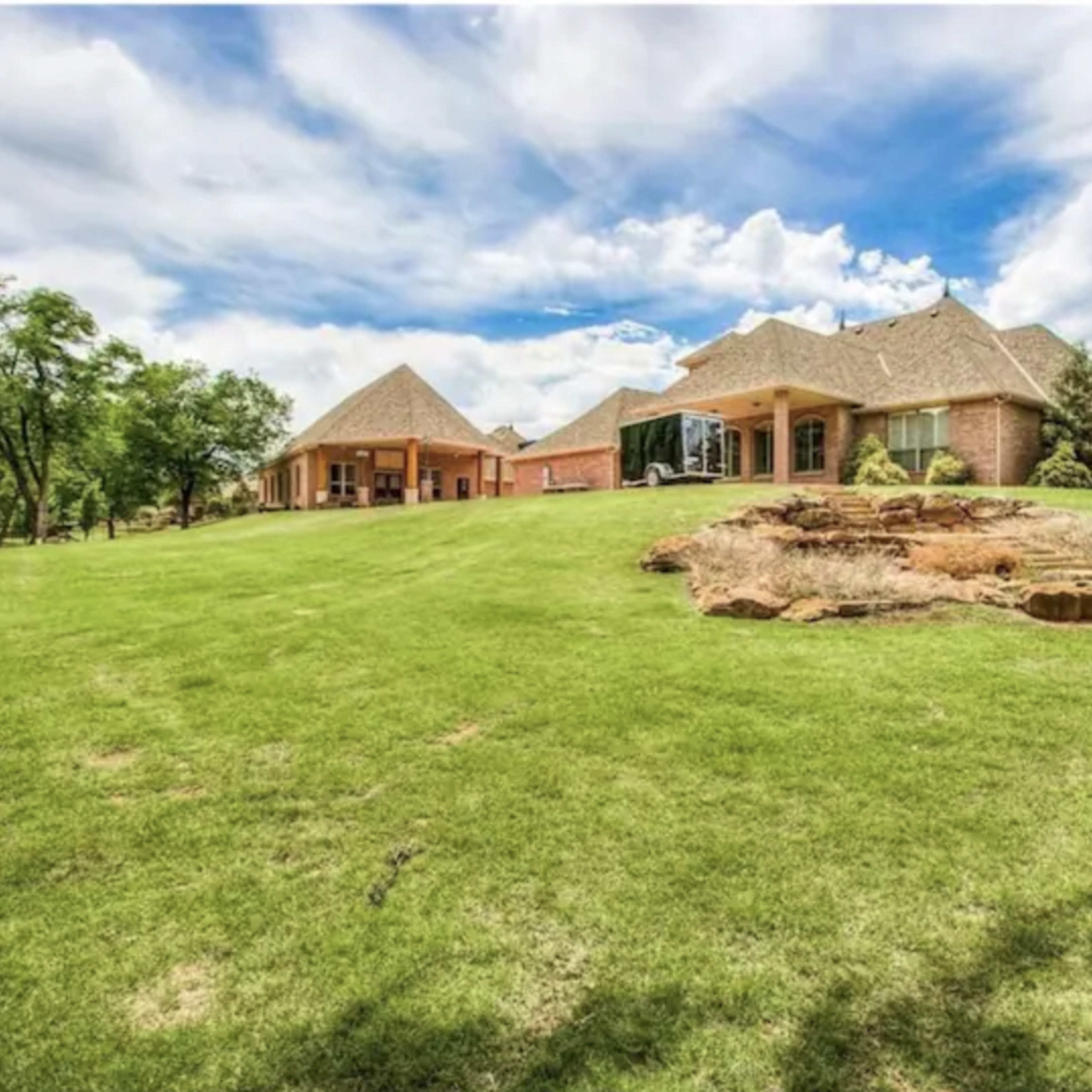 A large residential property features a brick house and a separate building on a lush green lawn under a partly cloudy sky.