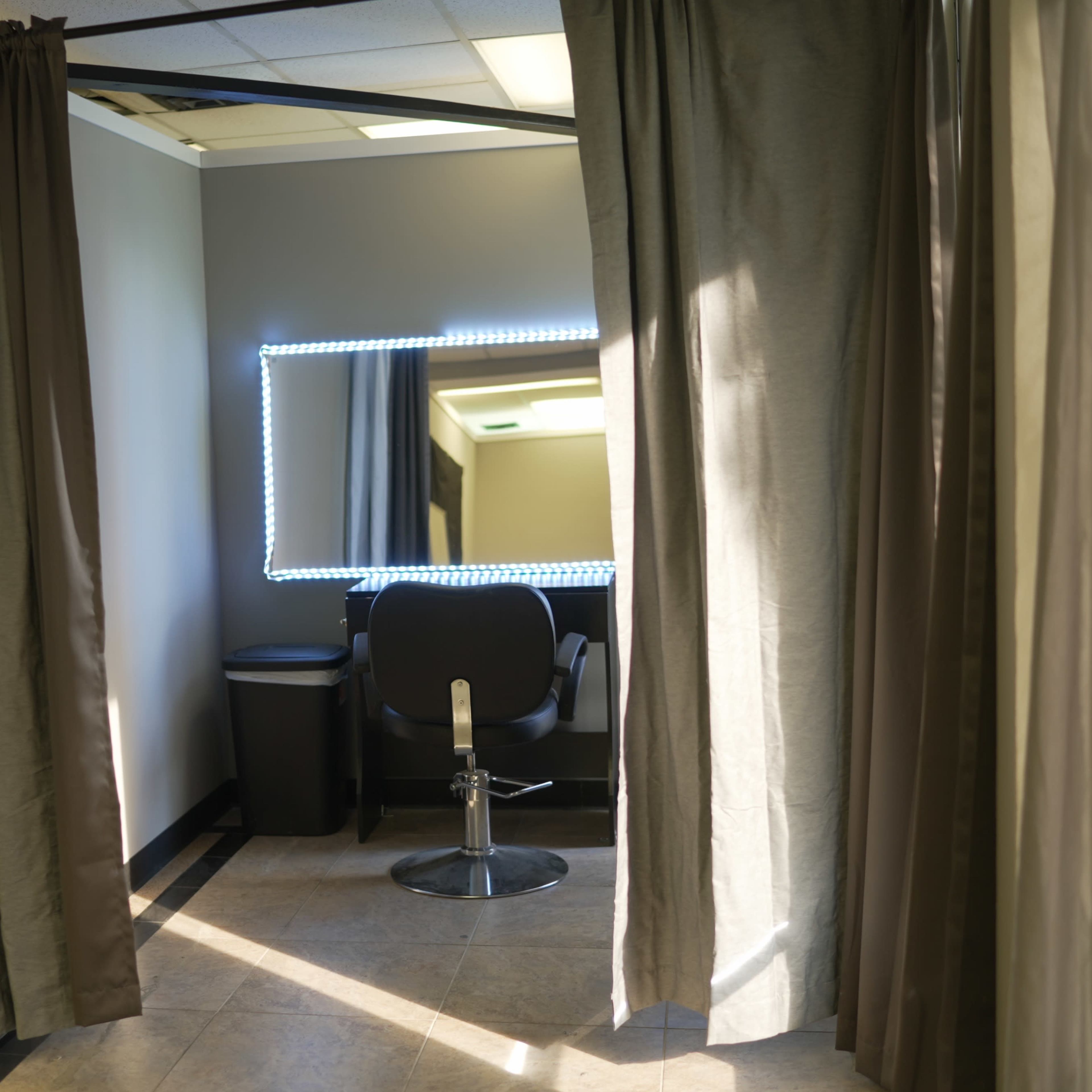 A salon space with a mirror surrounded by lights, a chair, and privacy curtains.