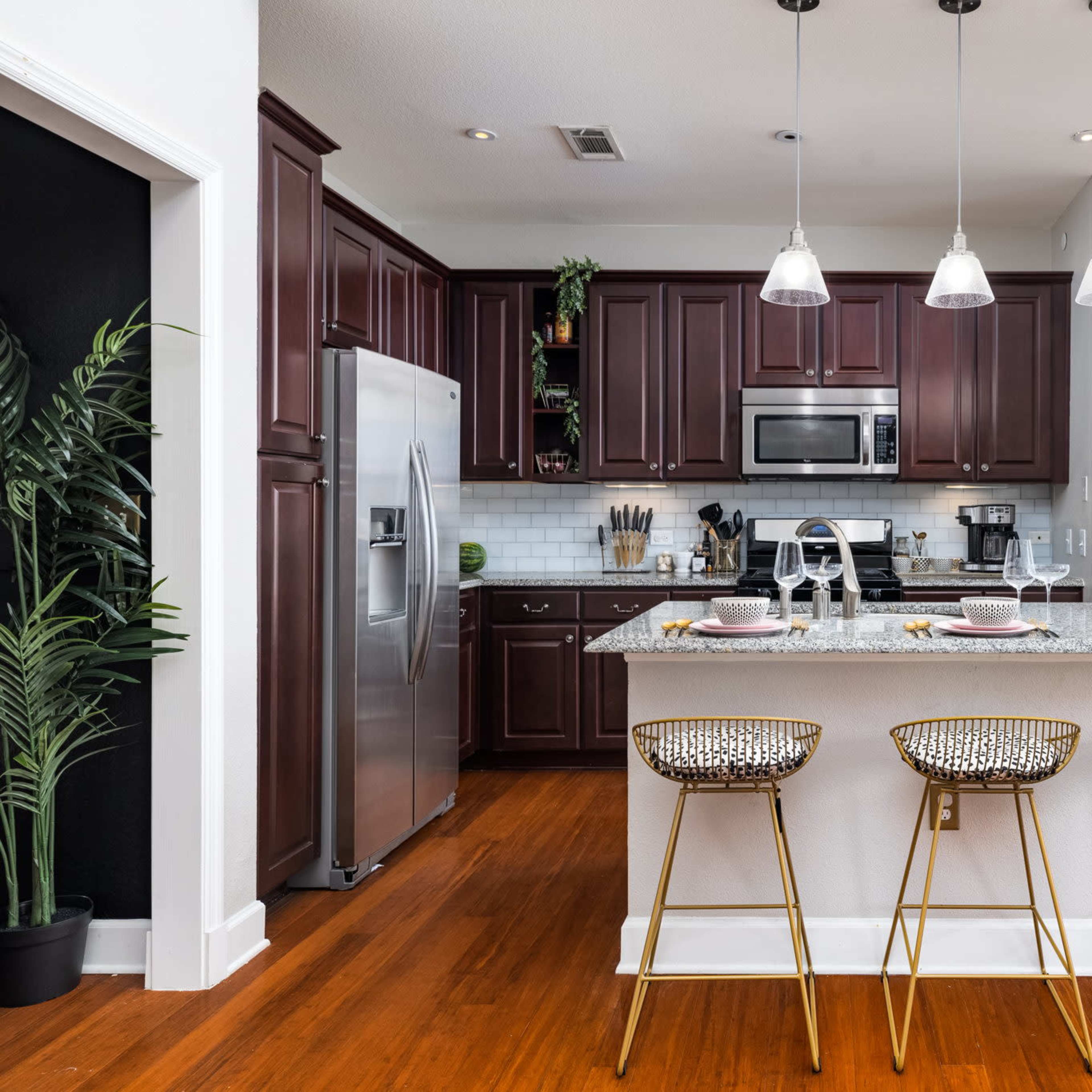 A modern kitchen features dark wood cabinetry, stainless steel appliances, and a granite island with three gold-accented bar stools.