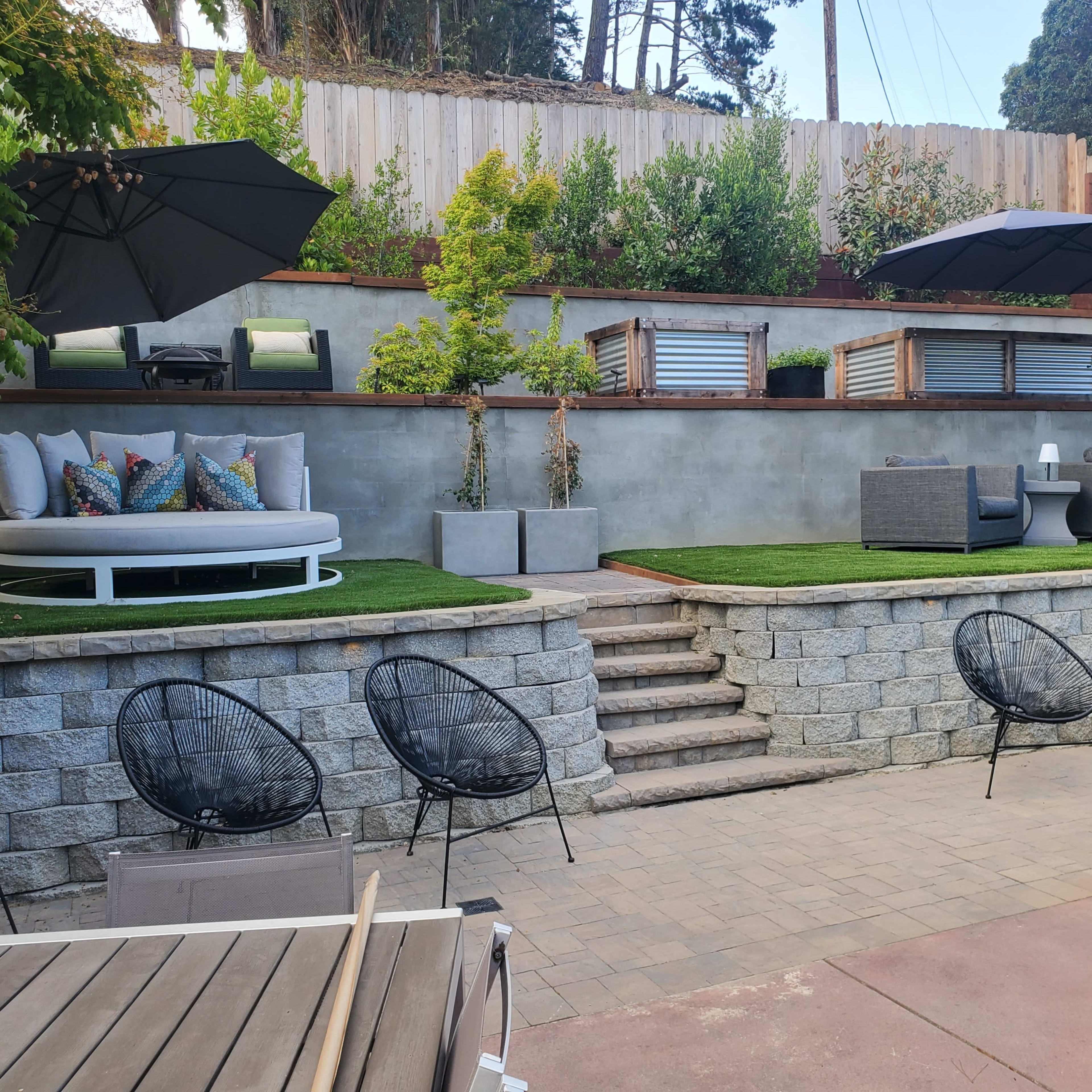 A landscaped backyard featuring tiered seating areas with outdoor furniture, umbrellas, and stone steps leading to a grassy space.