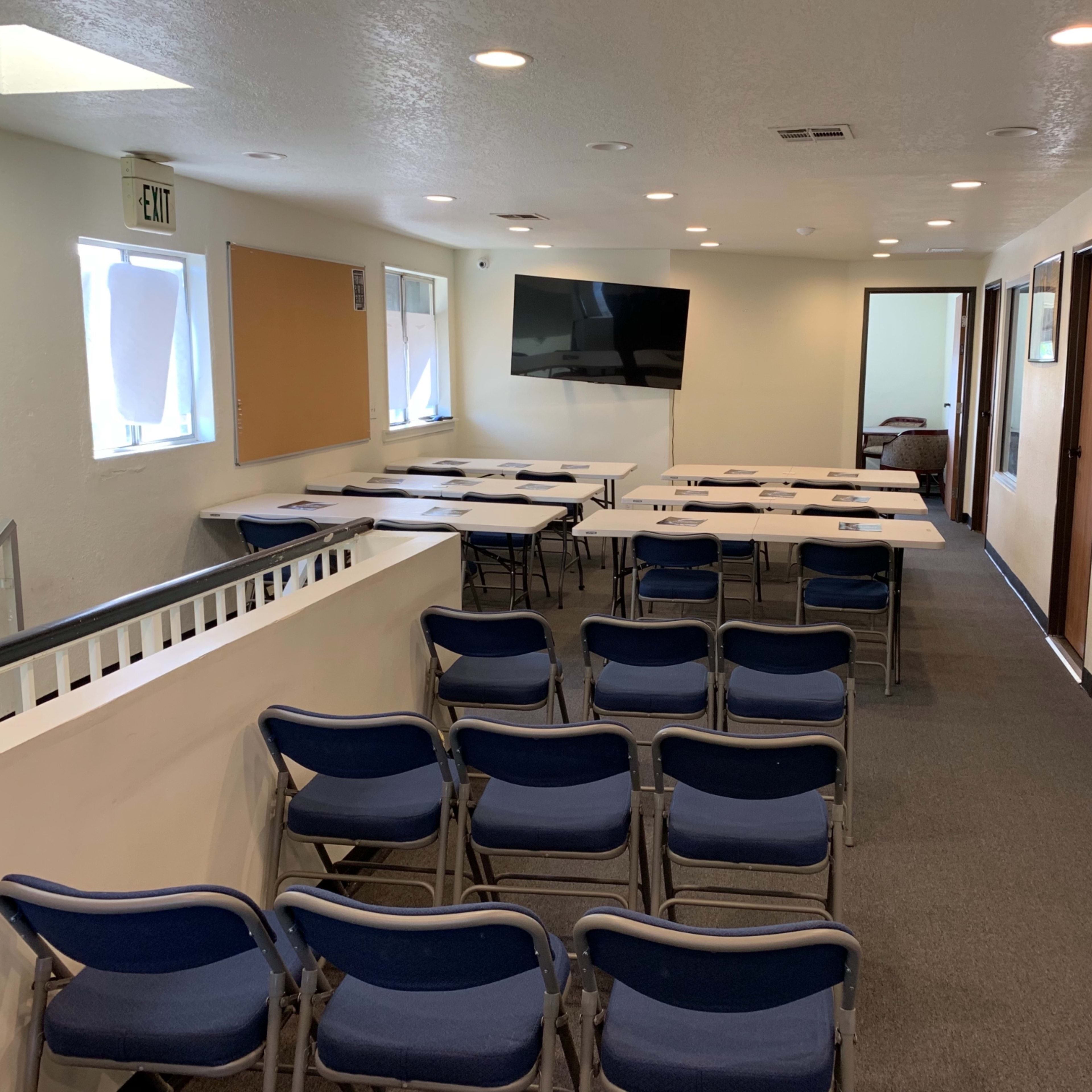 A meeting room arranged with rows of blue chairs facing a wall-mounted television, with tables set up for a presentation.