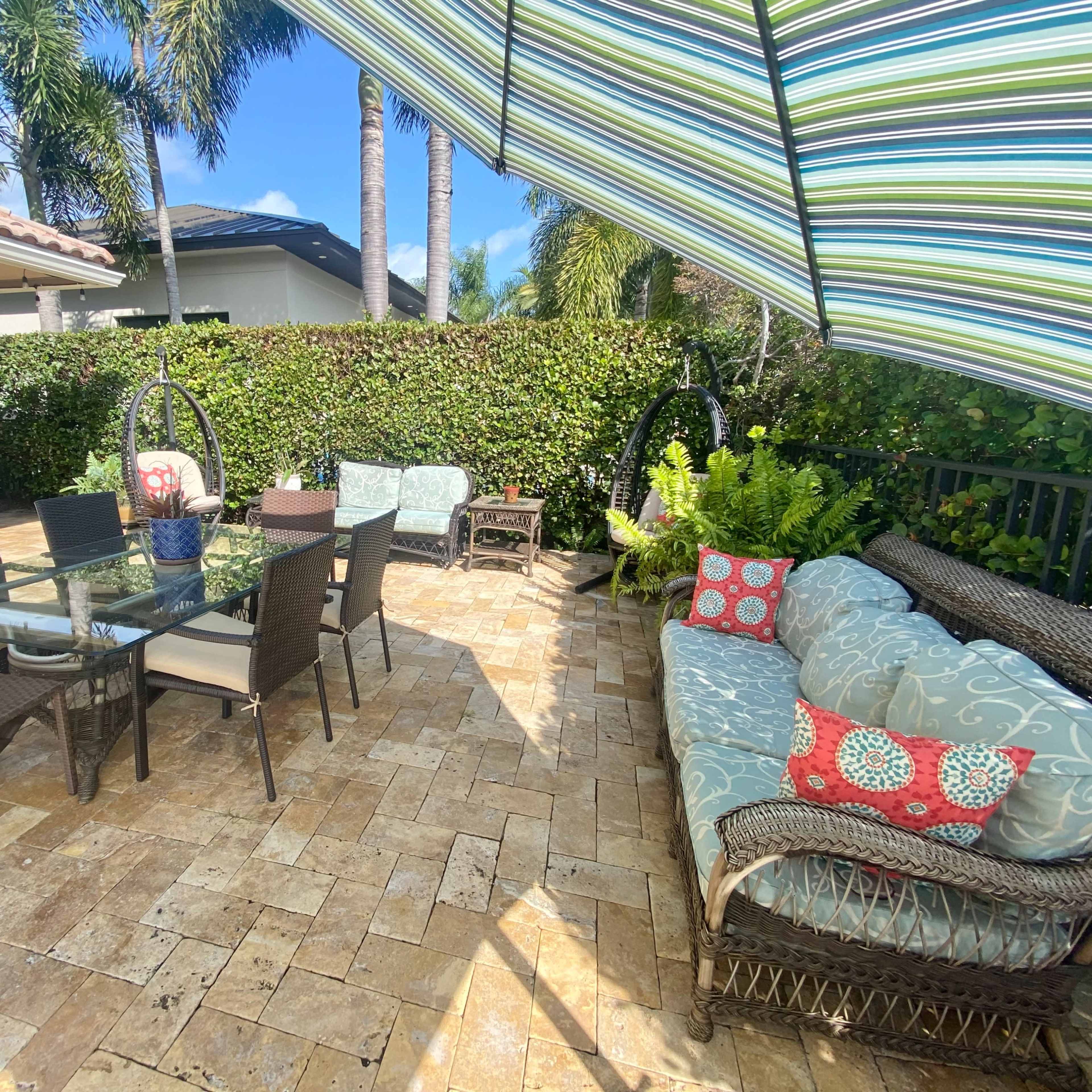 A patio area features a glass table surrounded by wicker chairs, seating with patterned cushions, and an umbrella for shade amidst lush greenery.