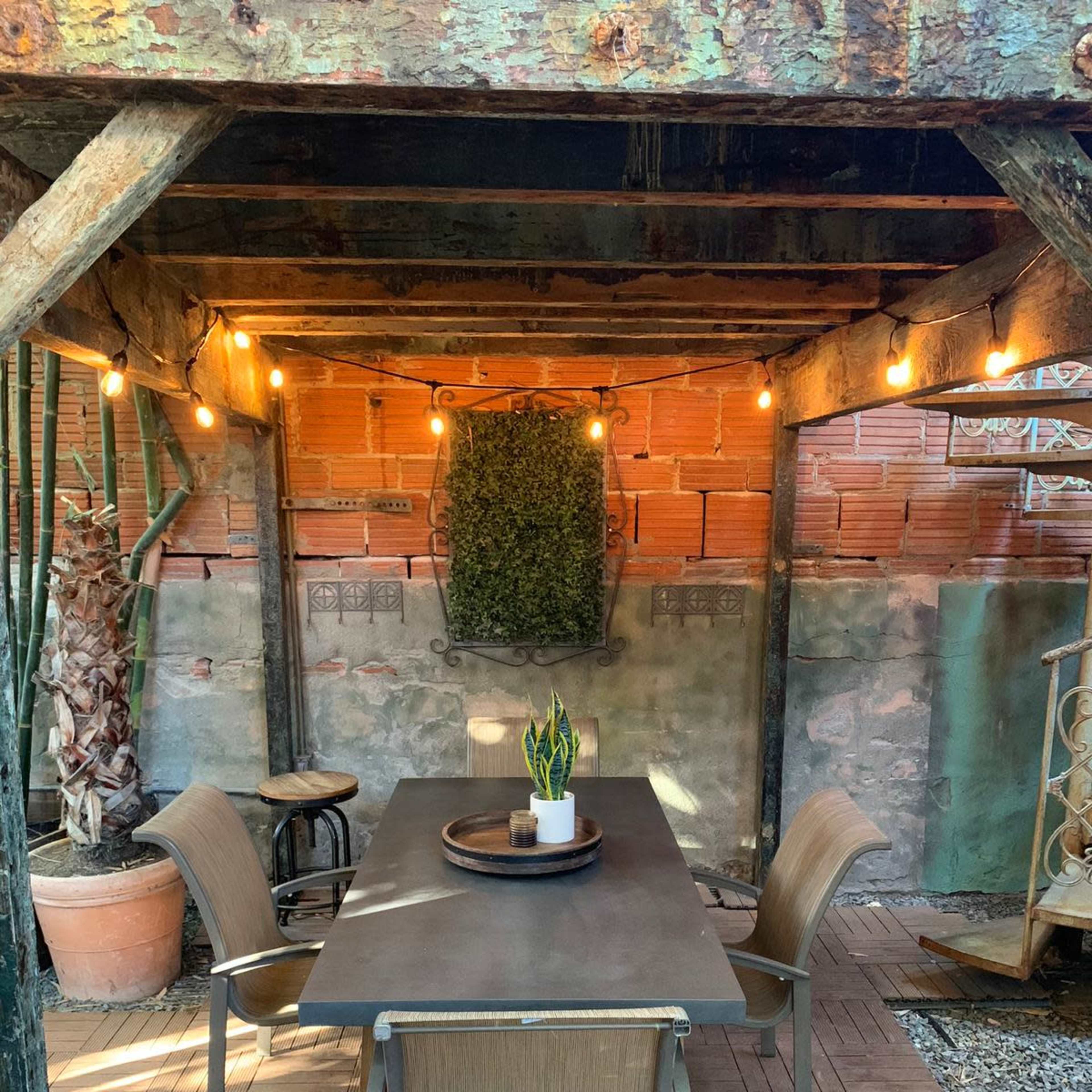 A wooden outdoor dining area features a rectangular table surrounded by four chairs, with string lights overhead and a hanging plant on the wall.