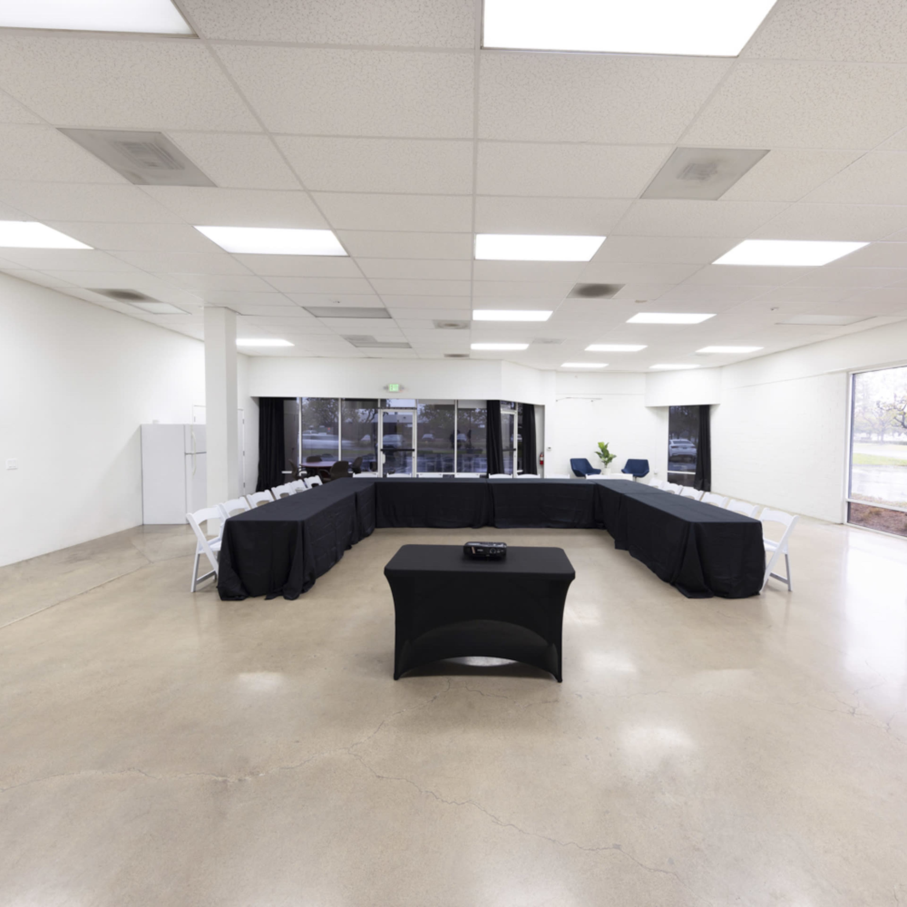 A large, empty conference room features a rectangular table covered with a black cloth, surrounded by chairs, and has large windows letting in natural light.