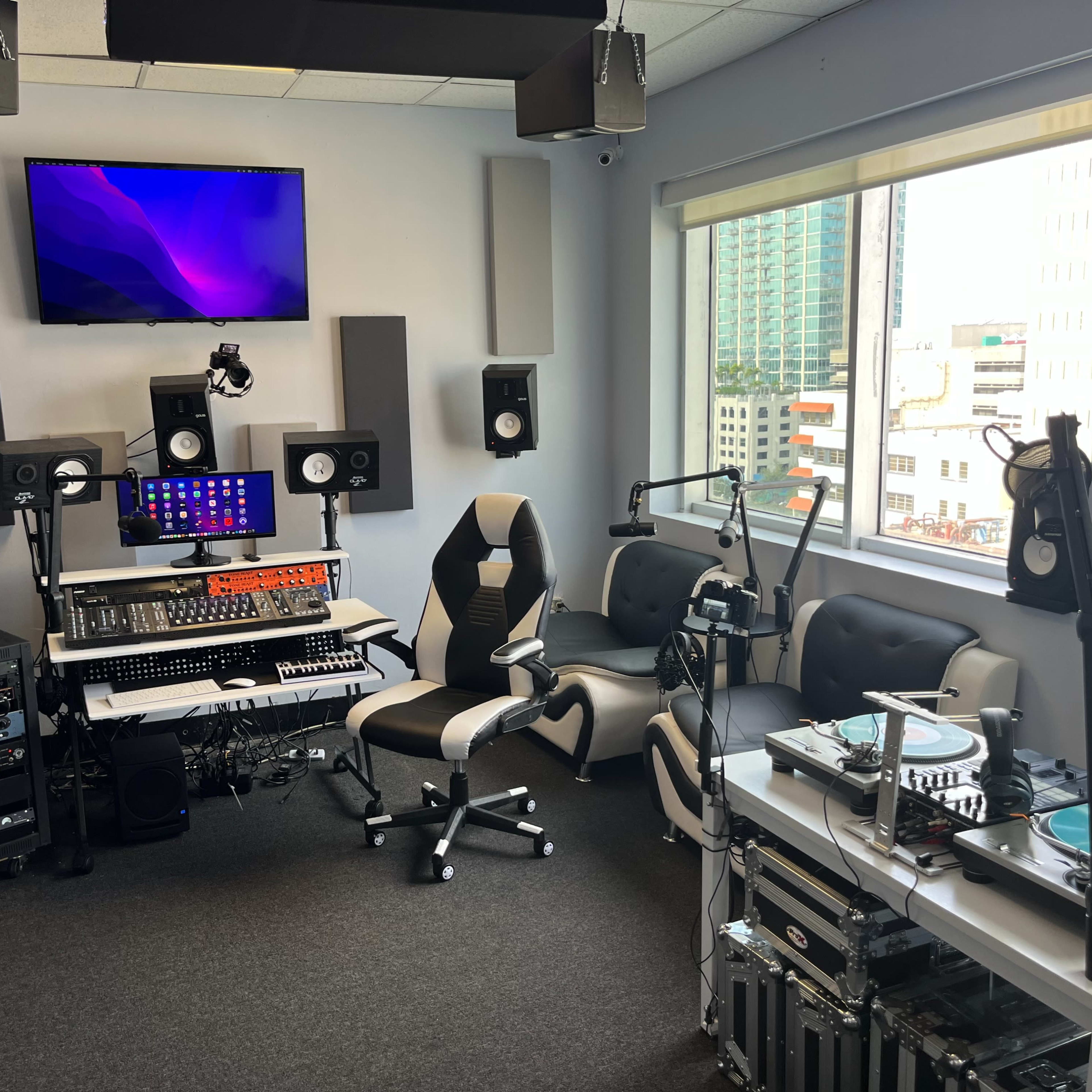 A modern music studio equipped with audio equipment, a computer setup, and seating near a large window overlooking a cityscape.