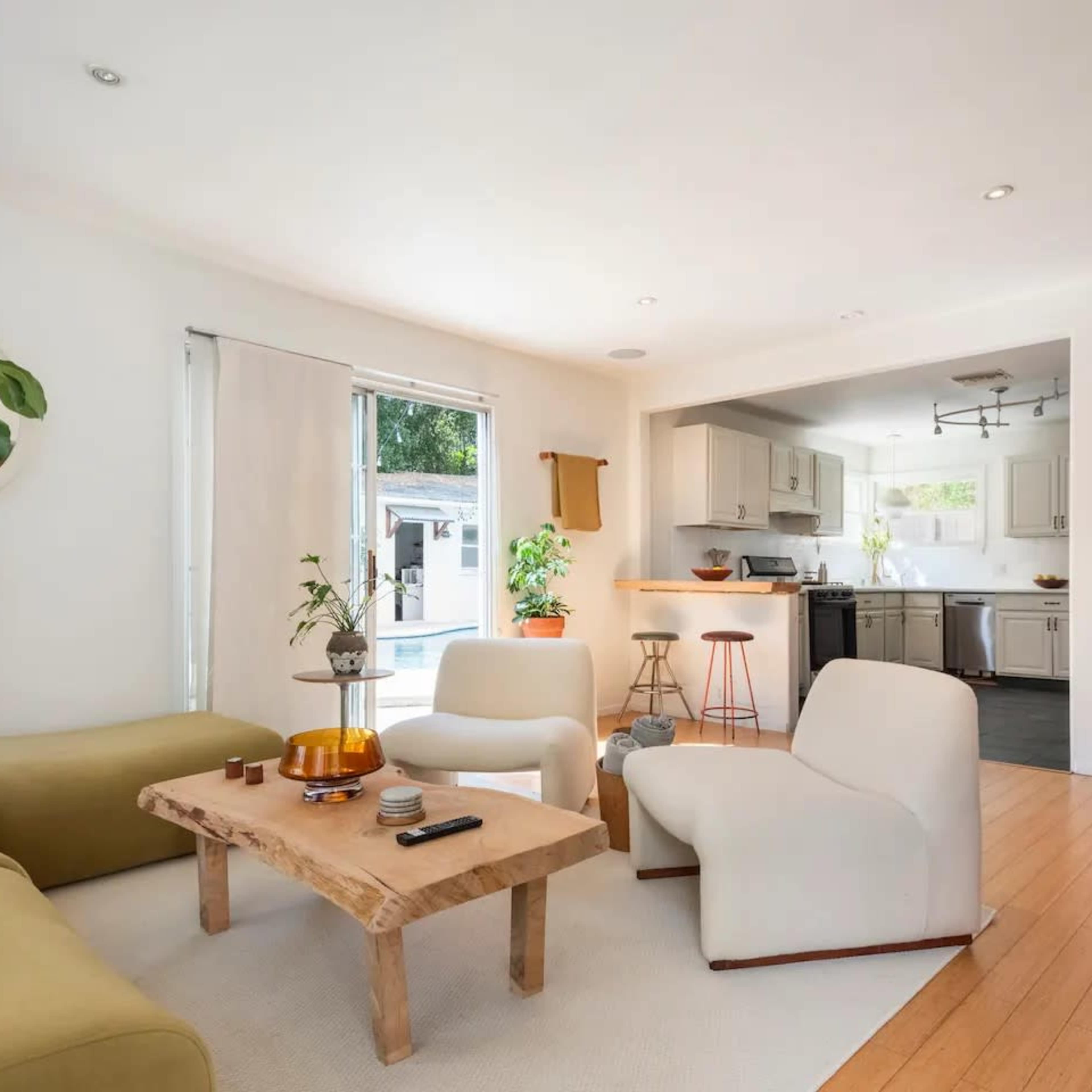 A modern living room with a green couch, white armchairs, a wooden coffee table, and an open kitchen area visible in the background.
