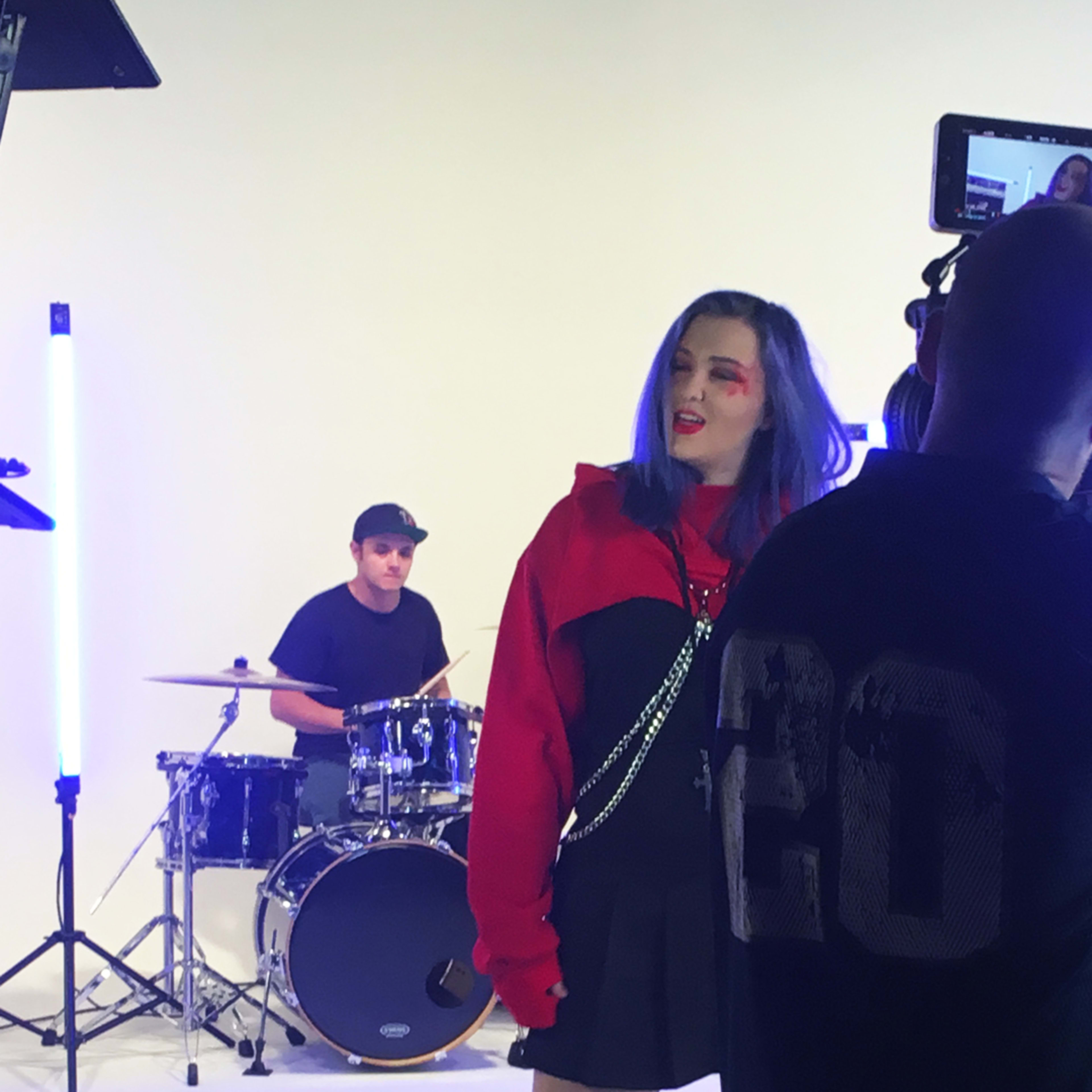 A musician in a red hoodie performs in front of a camera while a drummer plays in the background on a brightly lit set.