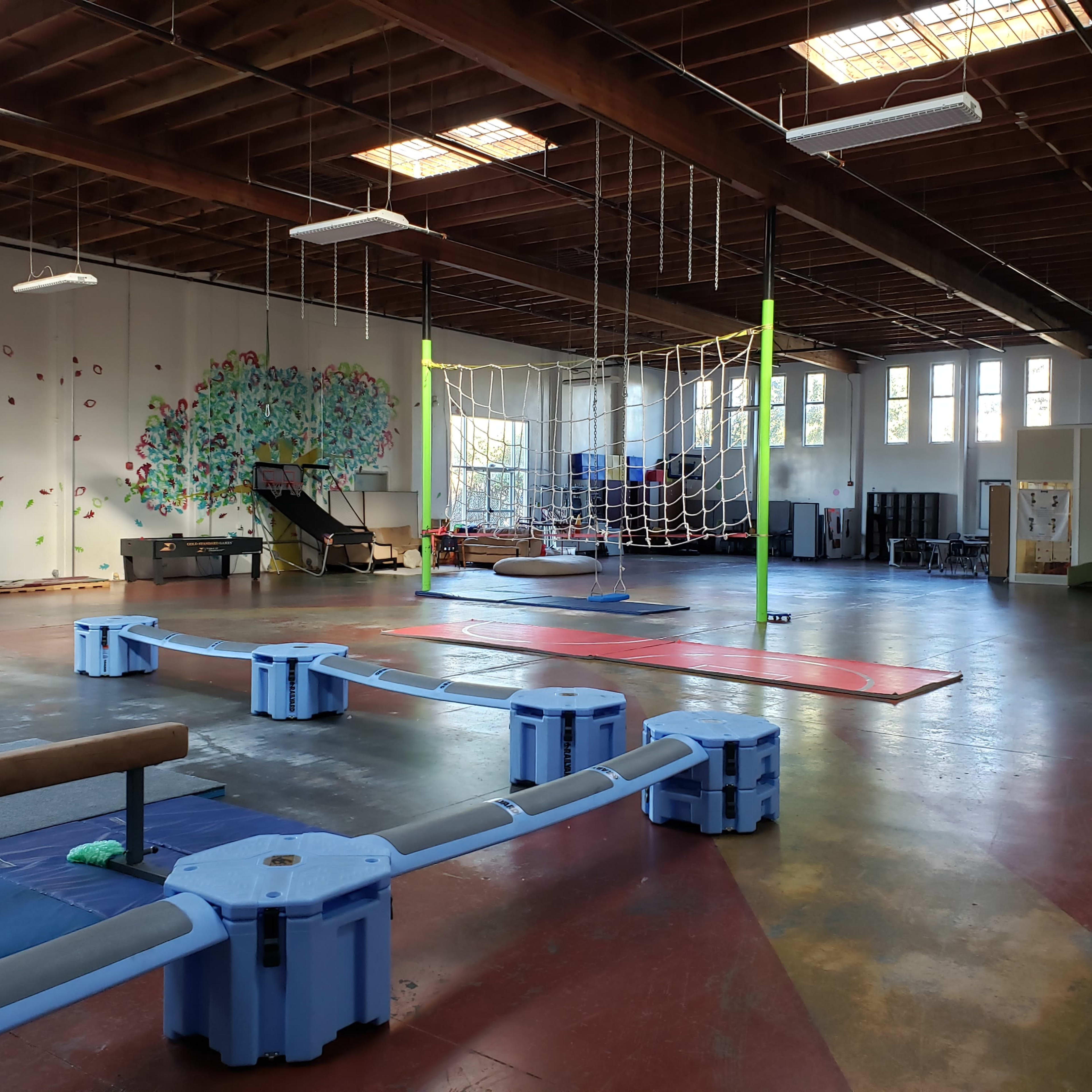 An indoor activity space with climbing structures, mats, and a rope net hanging from the ceiling.