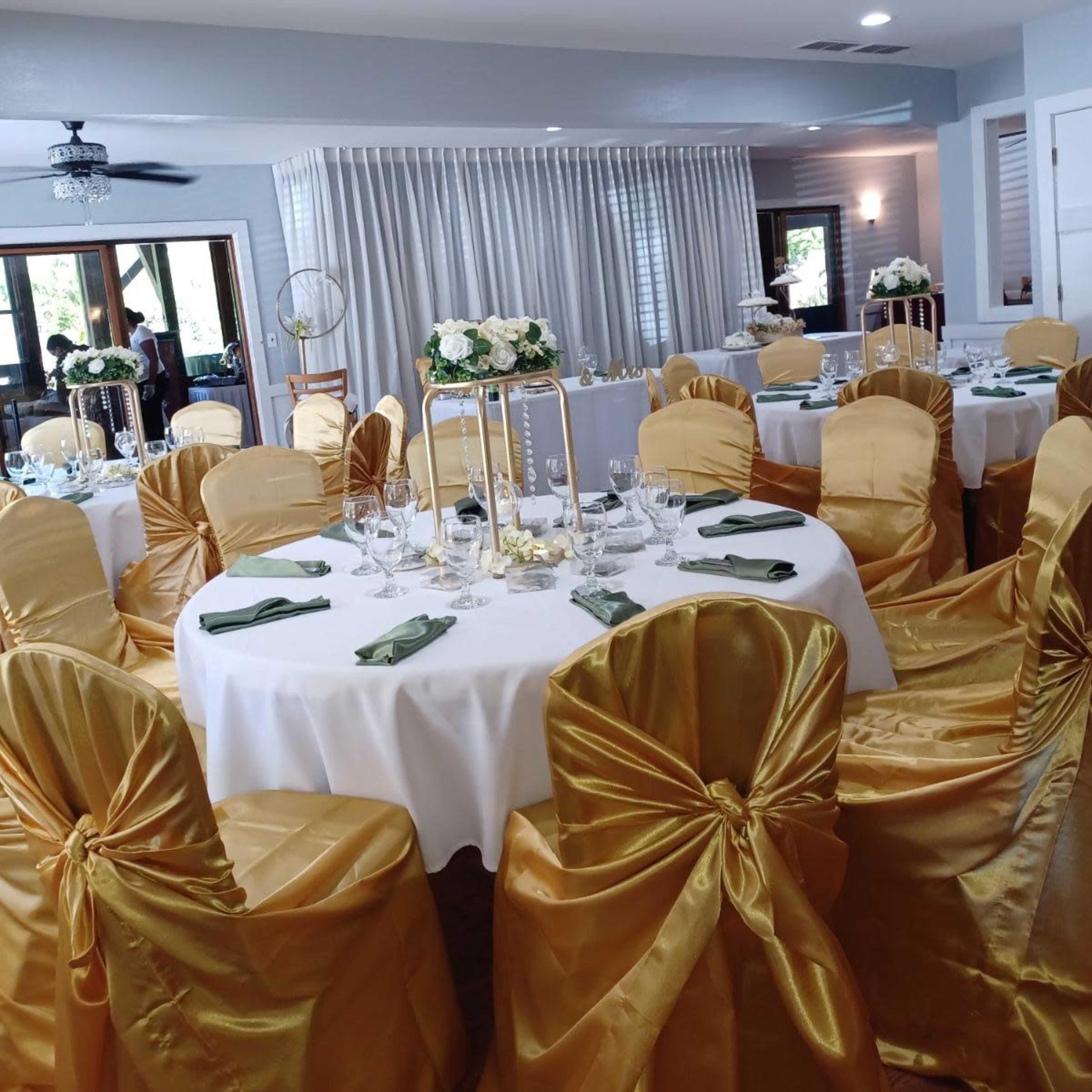 A dining area set for an event with round tables dressed in white tablecloths and finished with gold satin chair covers.