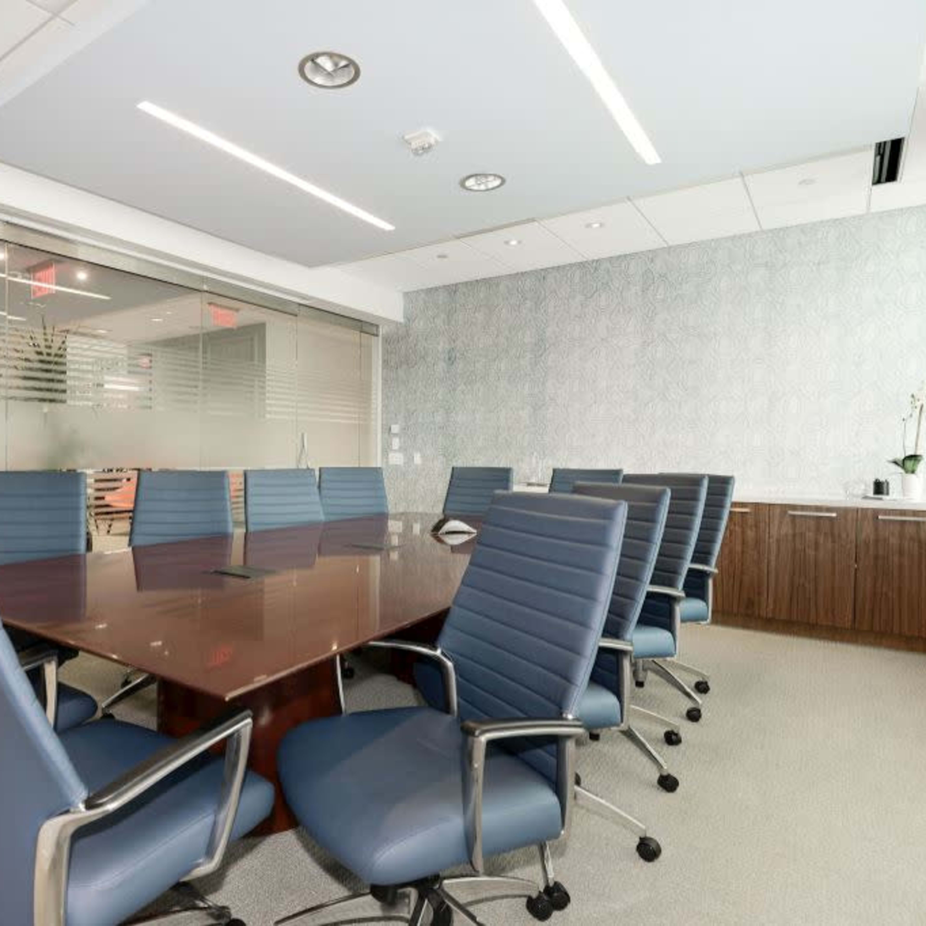 A modern conference room features a large wooden table surrounded by eight blue chairs, with glass walls and a decorative background.