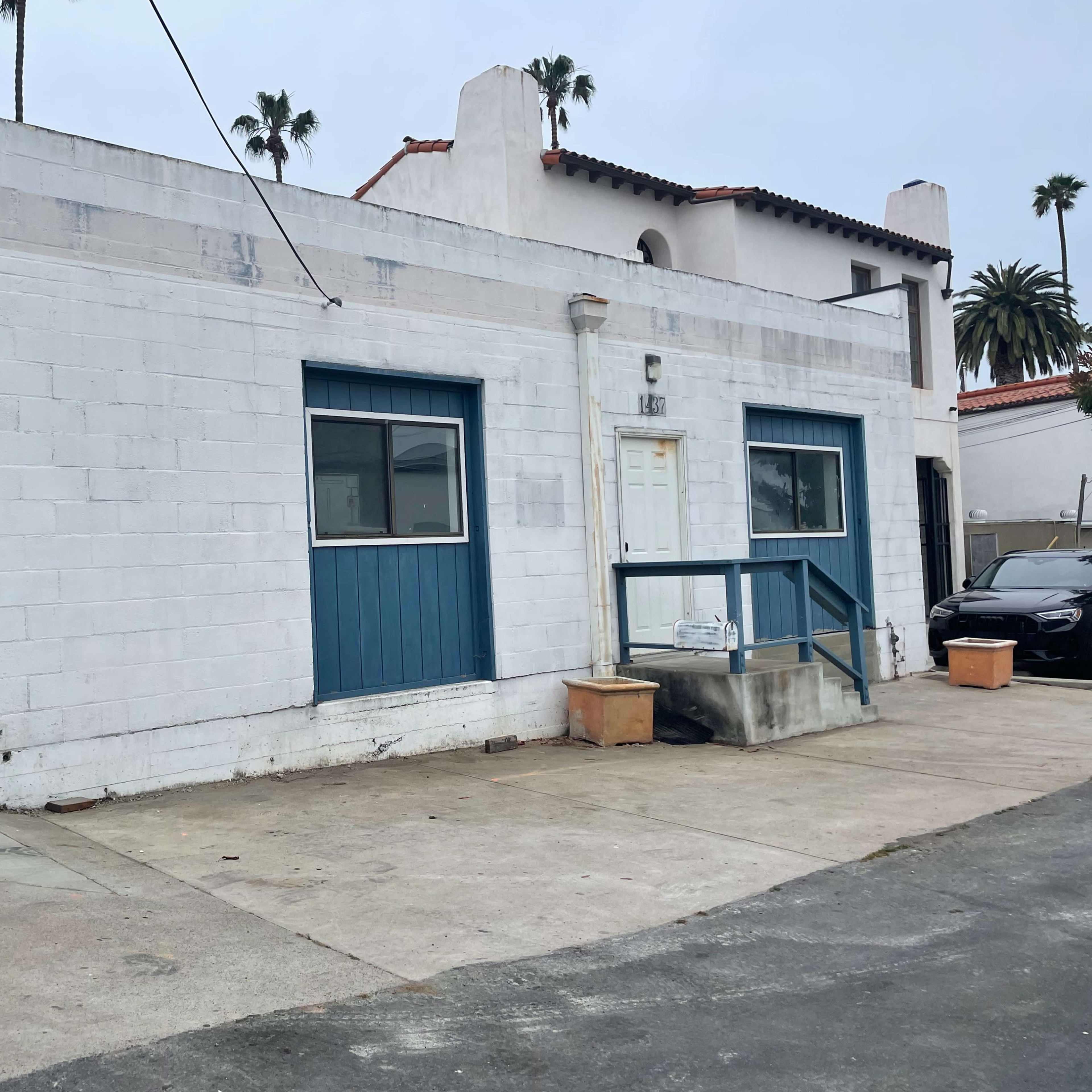 A white stucco building with a blue door and windows, set against a cloudy sky, surrounded by palm trees and pavement.