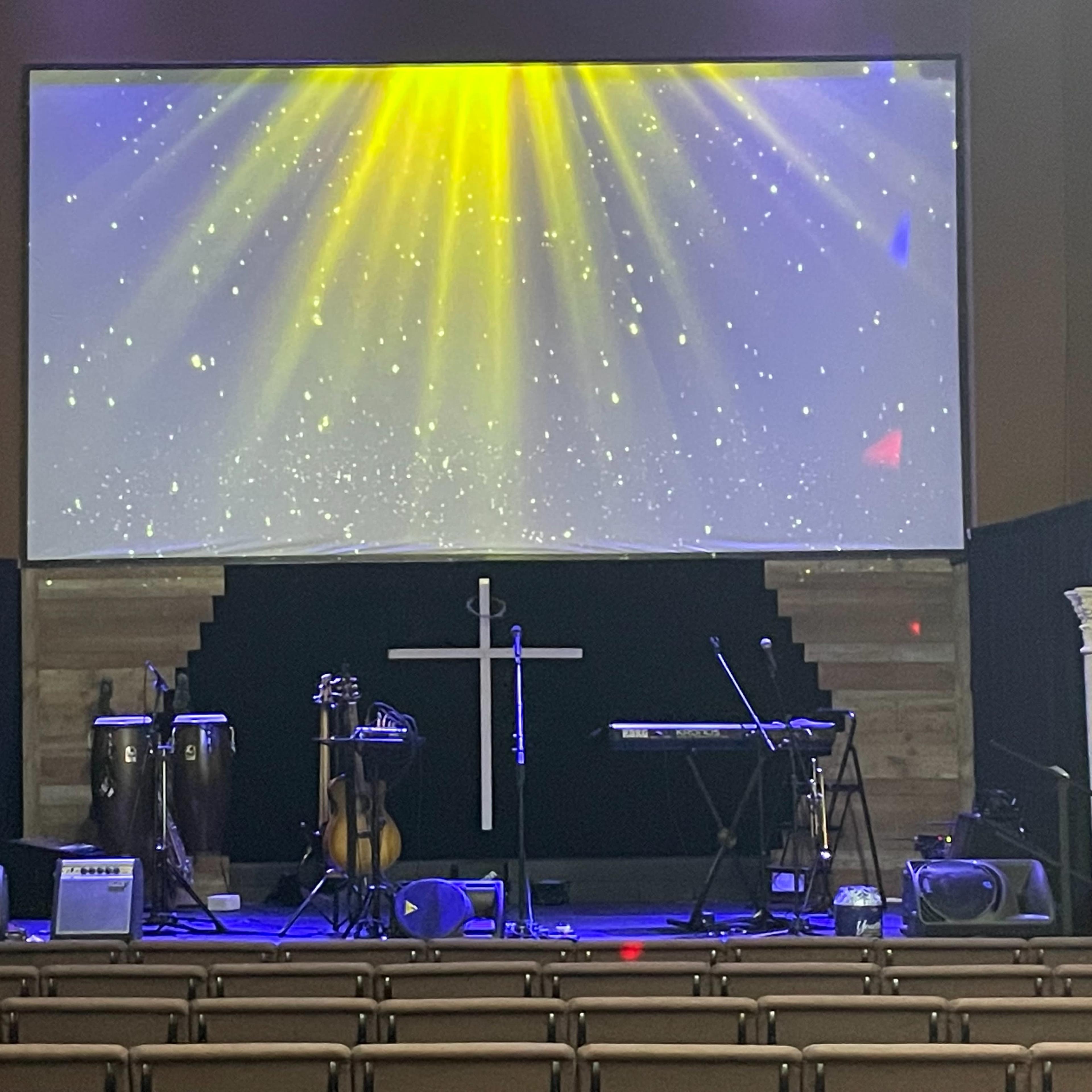 A stage in a church setting, featuring musical instruments like congas and a keyboard, with a large screen displaying beams of light in the background.