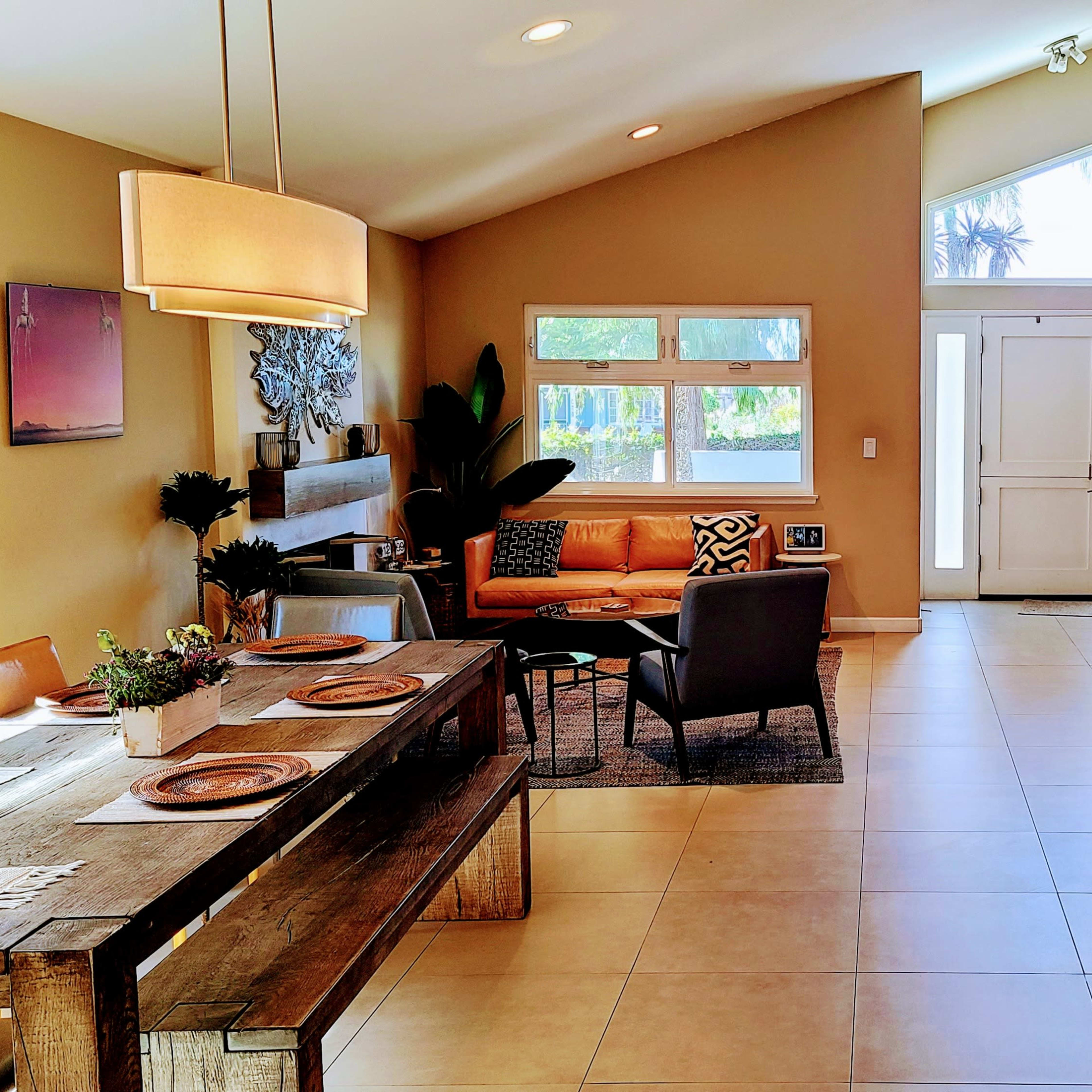 A modern, open-concept living space featuring a dining area with a wooden table, a cozy sitting area with a couch, and an entrance with large windows and a staircase.