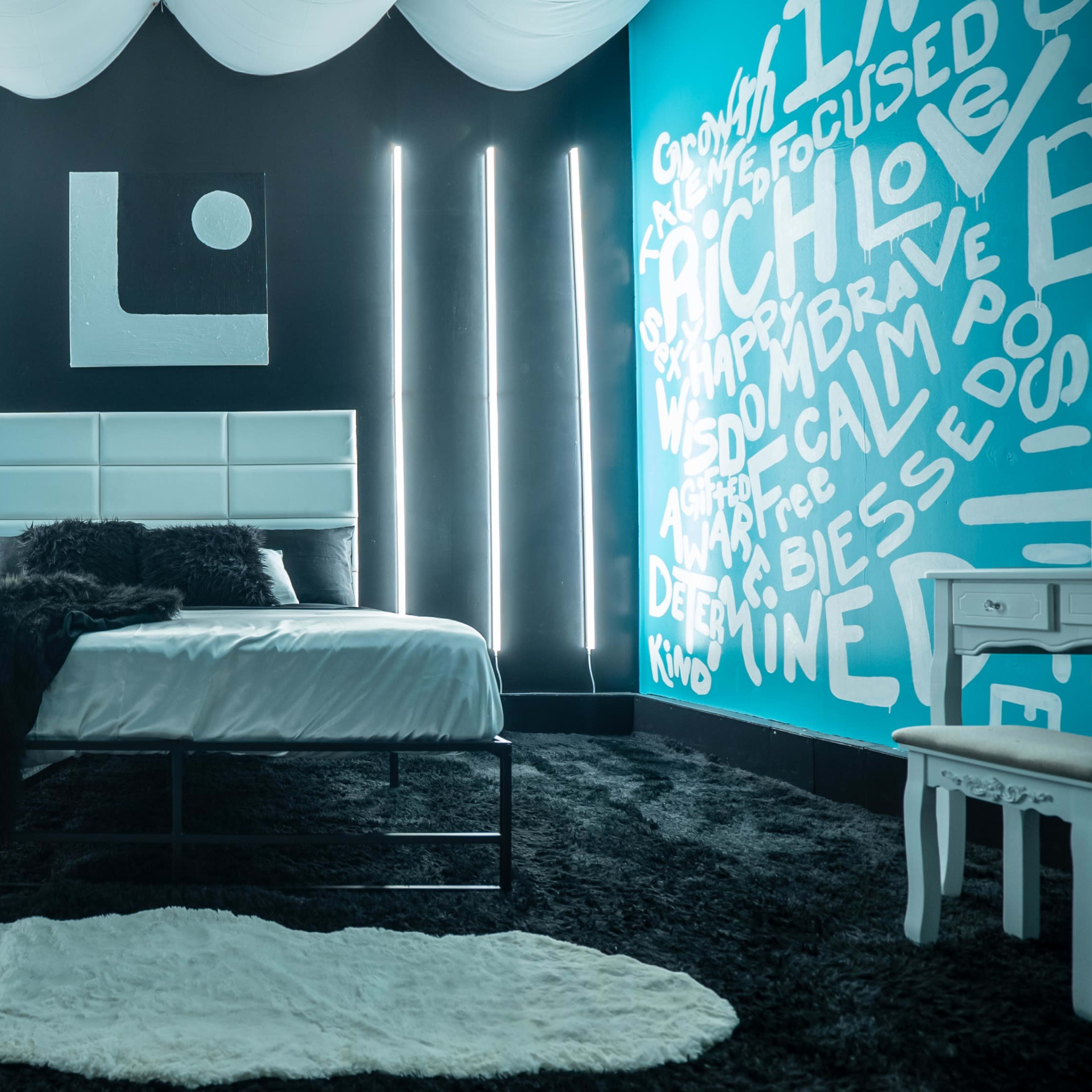 A modern bedroom with a bed covered in white linens, a black shag carpet, and walls painted in contrasting teal and black, featuring decorative lighting and text art.