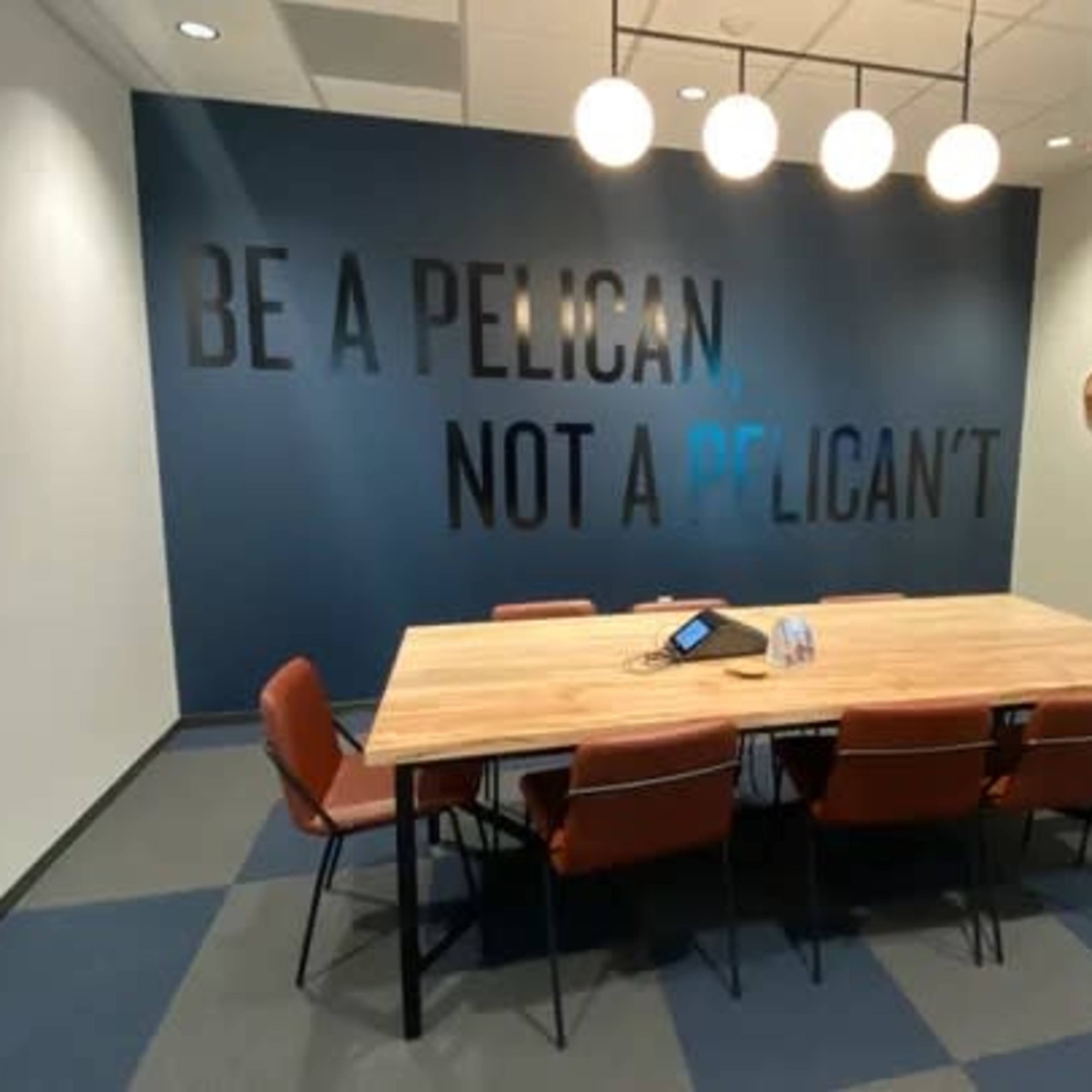 A conference room features a large wooden table surrounded by chairs, with a wall displaying the phrase "BE A PELICAN NOT A PELICAN'T" and a screen mounted on one side.