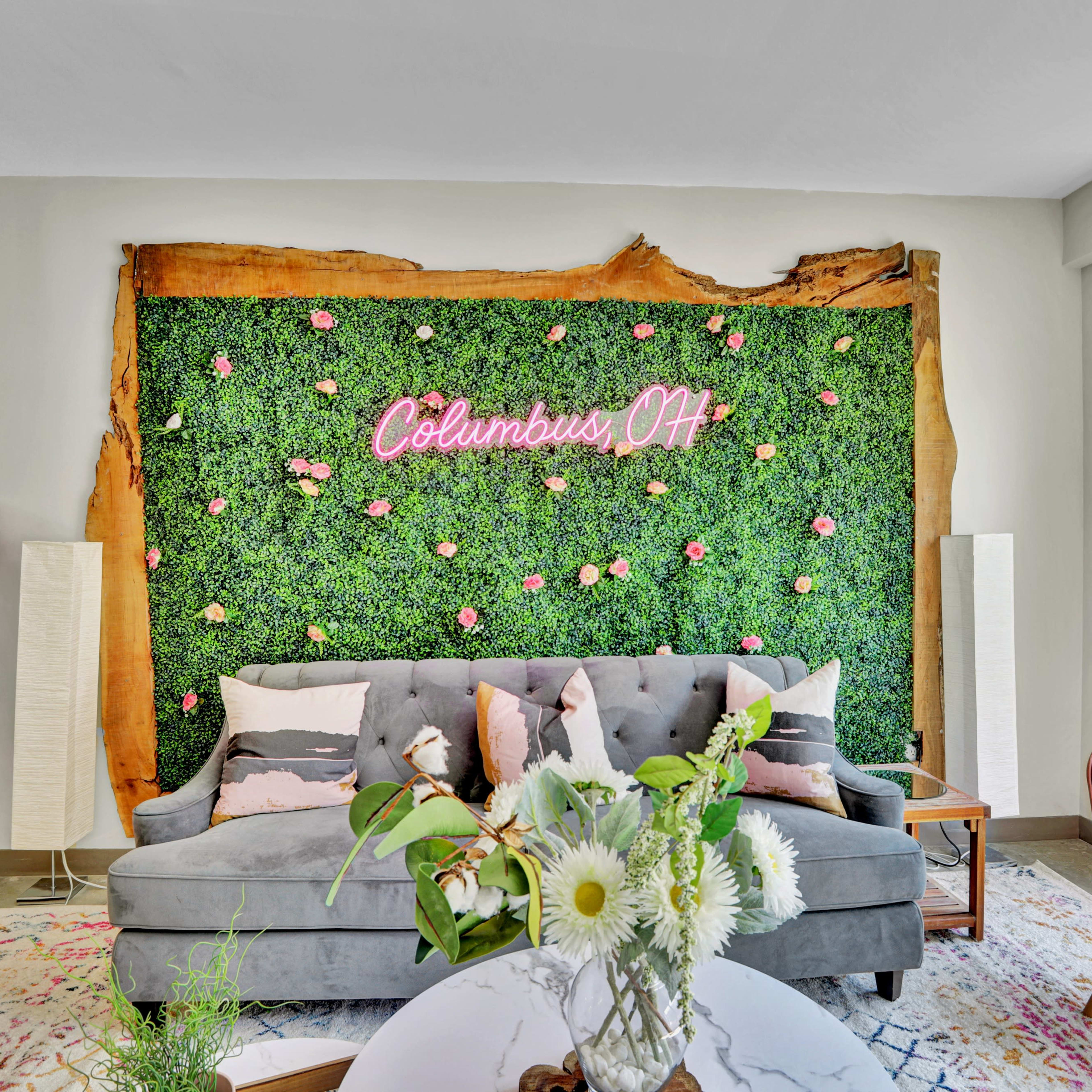 A living room with a gray sofa in front of a green wall adorned with pink flowers and a neon sign that reads "Columbus, OH."