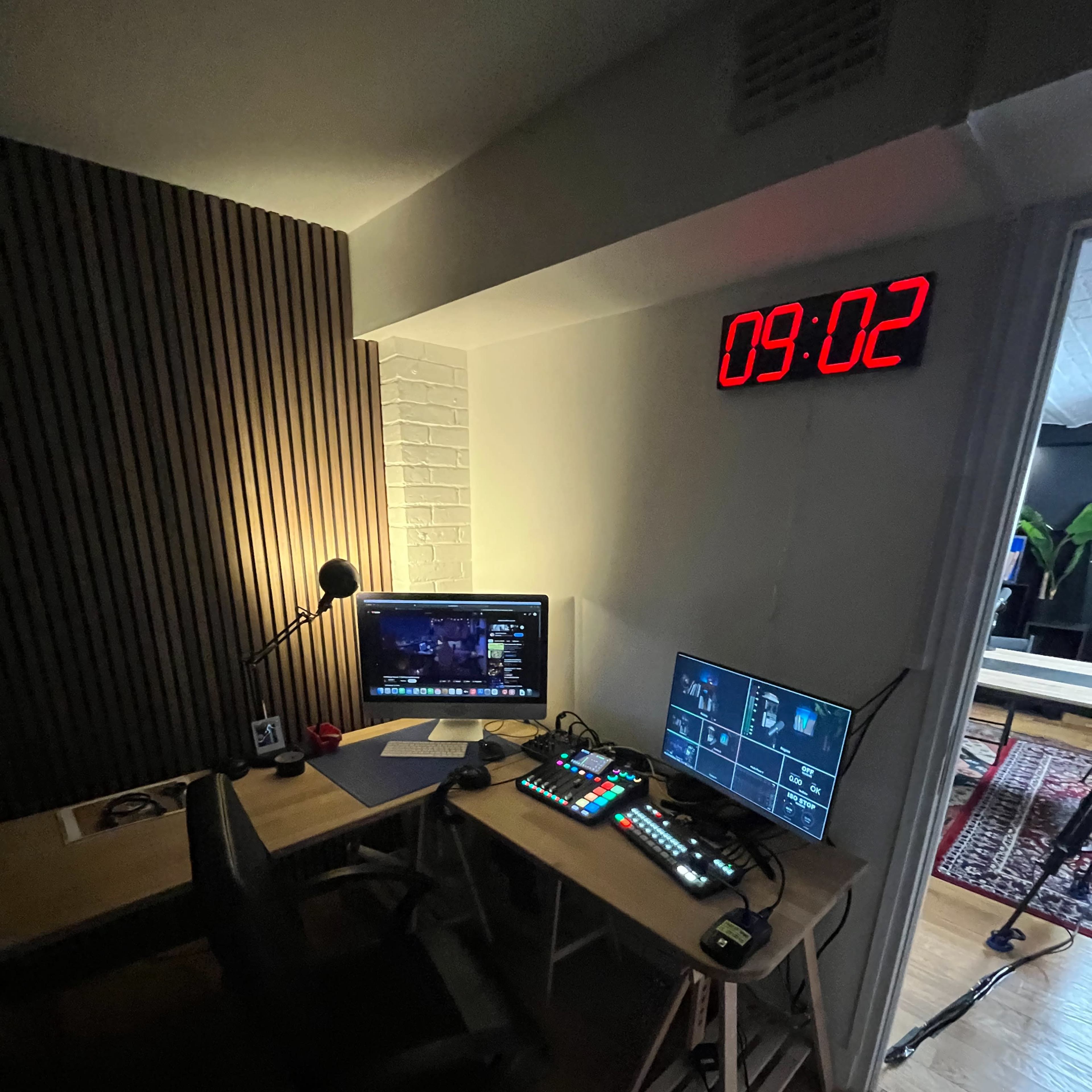 A modern workspace featuring a desk with a computer monitor, a second screen displaying controls, and a large digital clock on the wall.