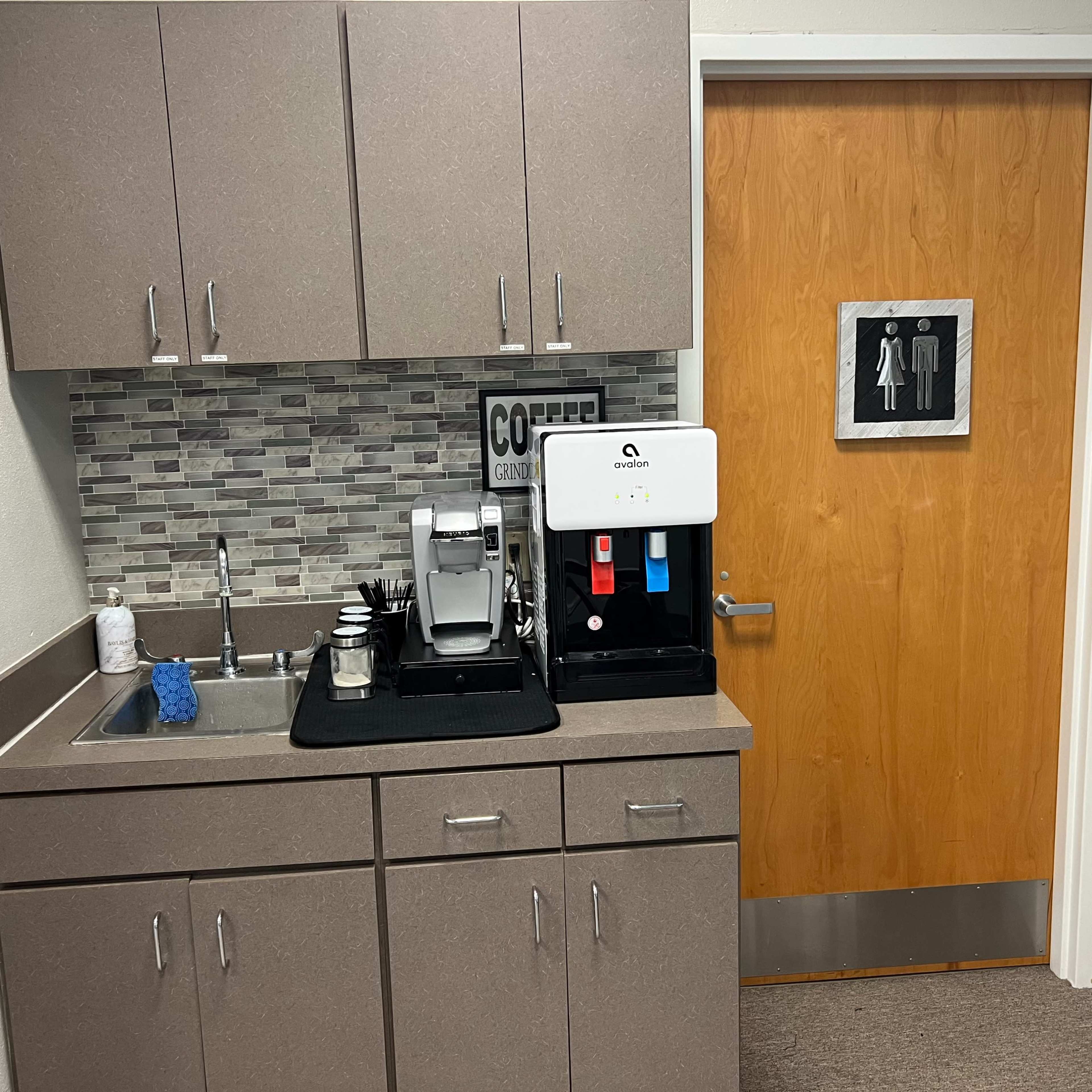 A small kitchenette area with a coffee machine and sink next to a bathroom door with gendered signage.