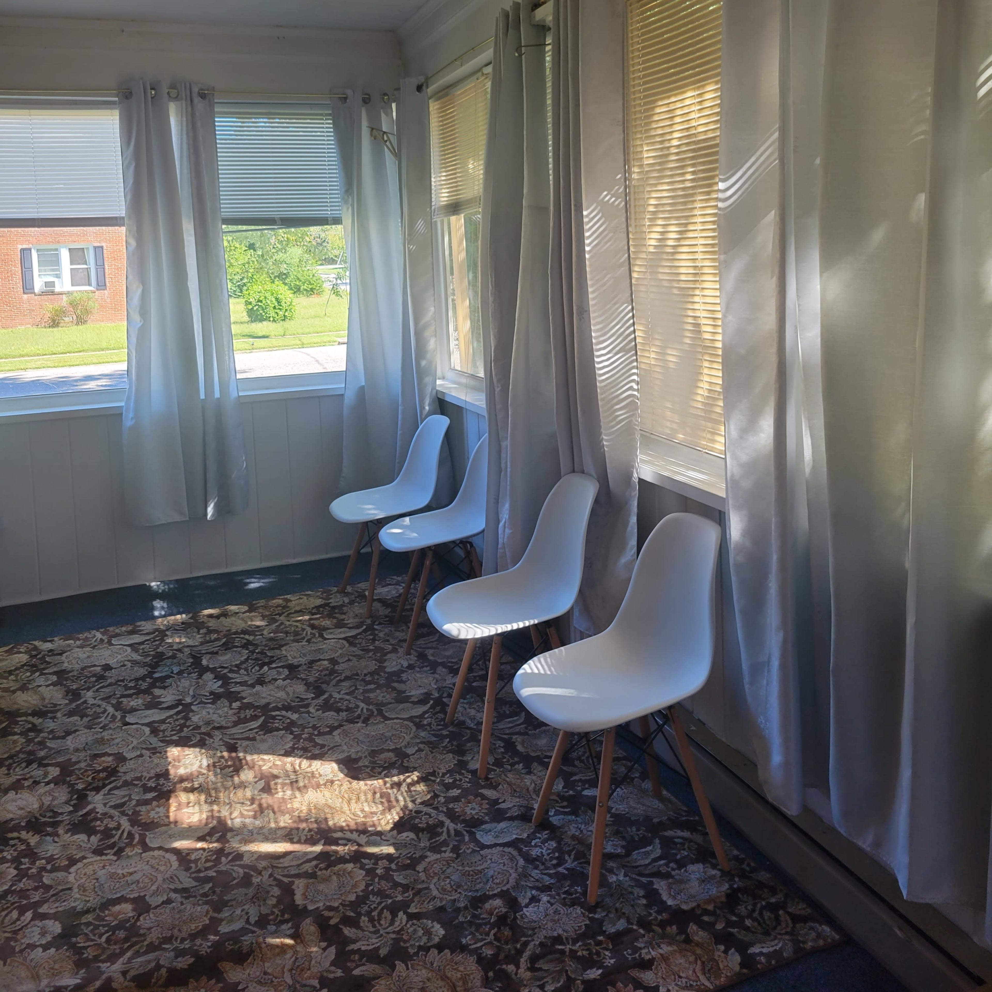 A sunlit, enclosed porch with three white chairs, large windows draped in sheer curtains, and a patterned rug on the floor.