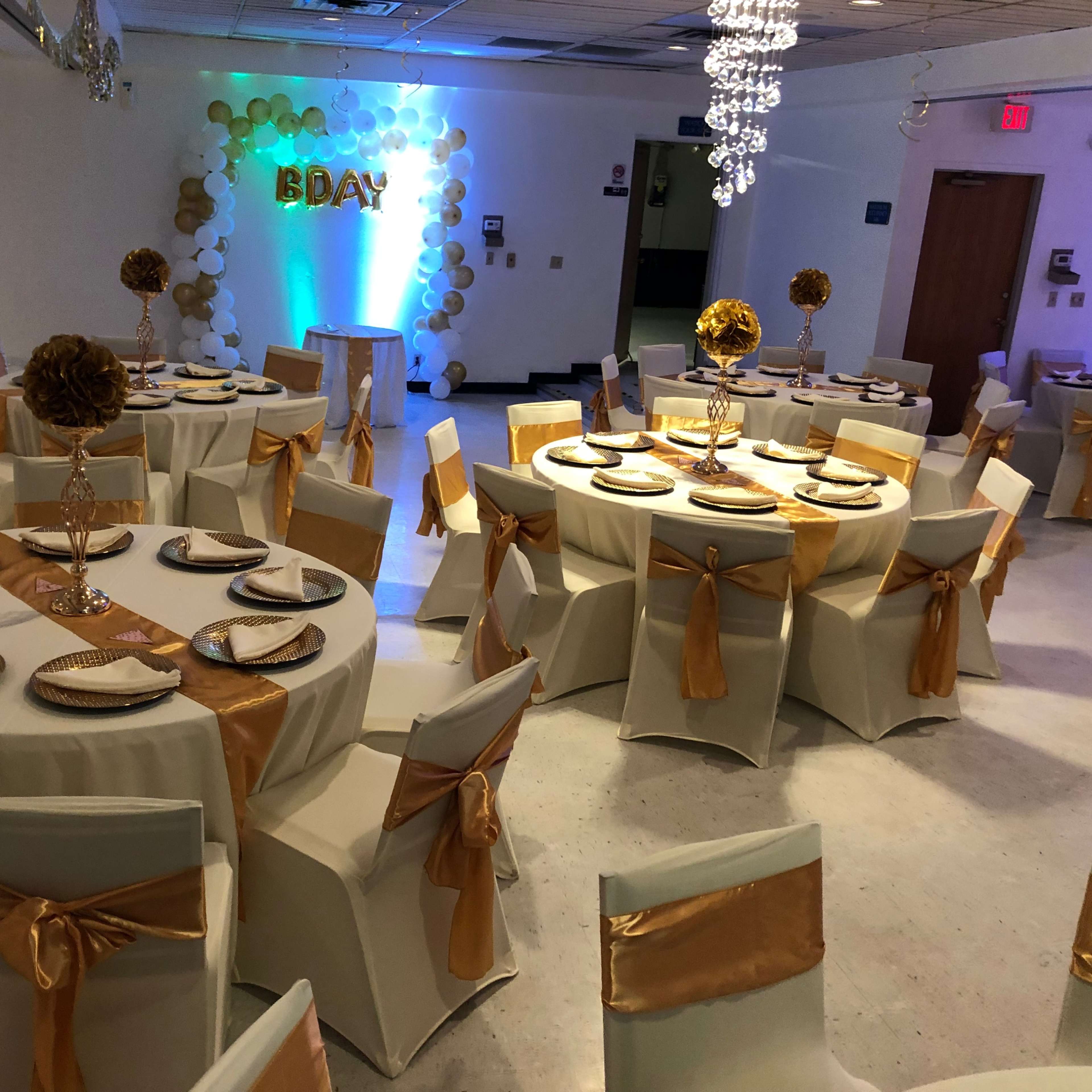 A decorated indoor event space with round tables covered in white tablecloths, gold accents, and a backdrop displaying "BDAY" surrounded by balloons.