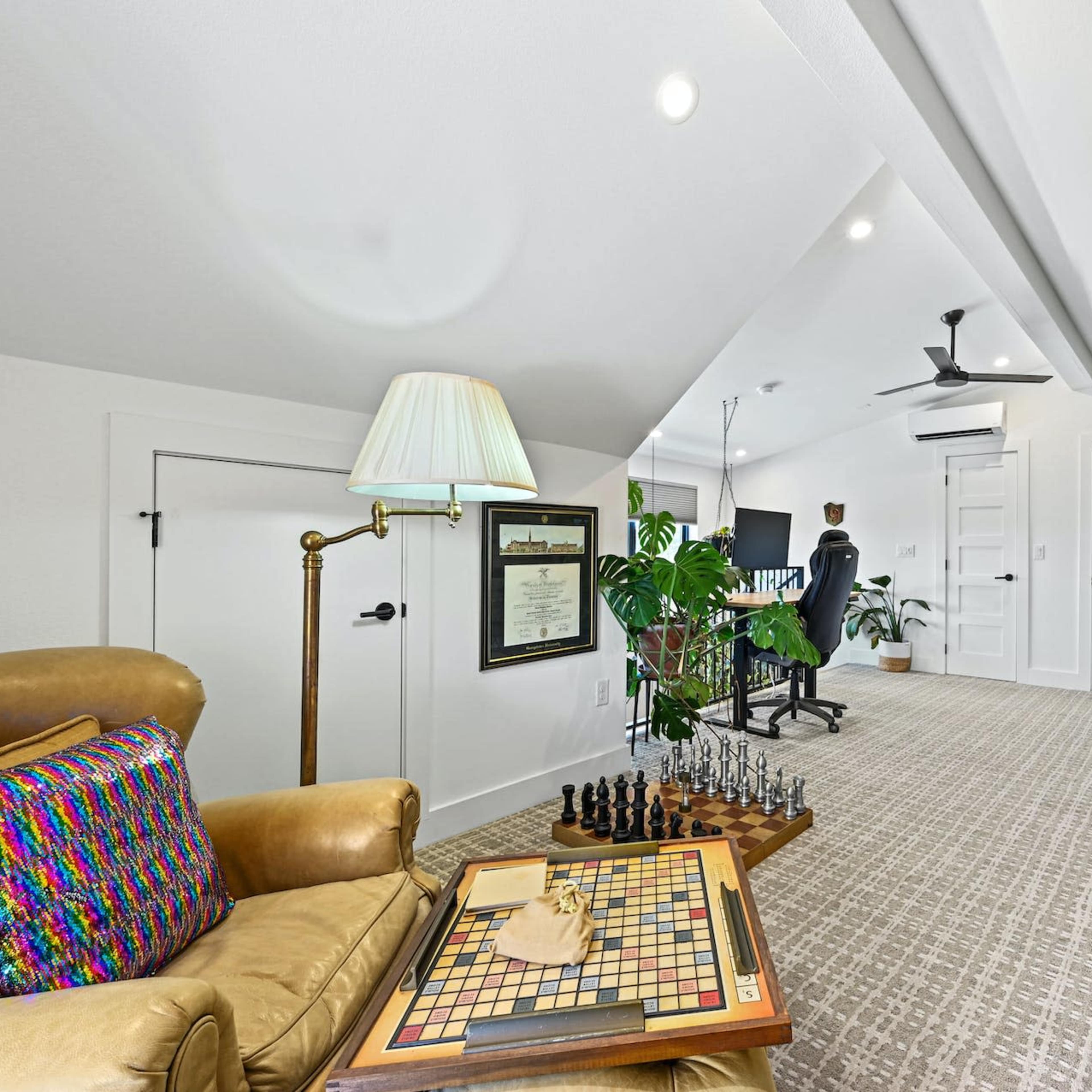 A spacious, bright attic room furnished with a comfortable chair, a chess set, a desk with a computer, and several green plants.