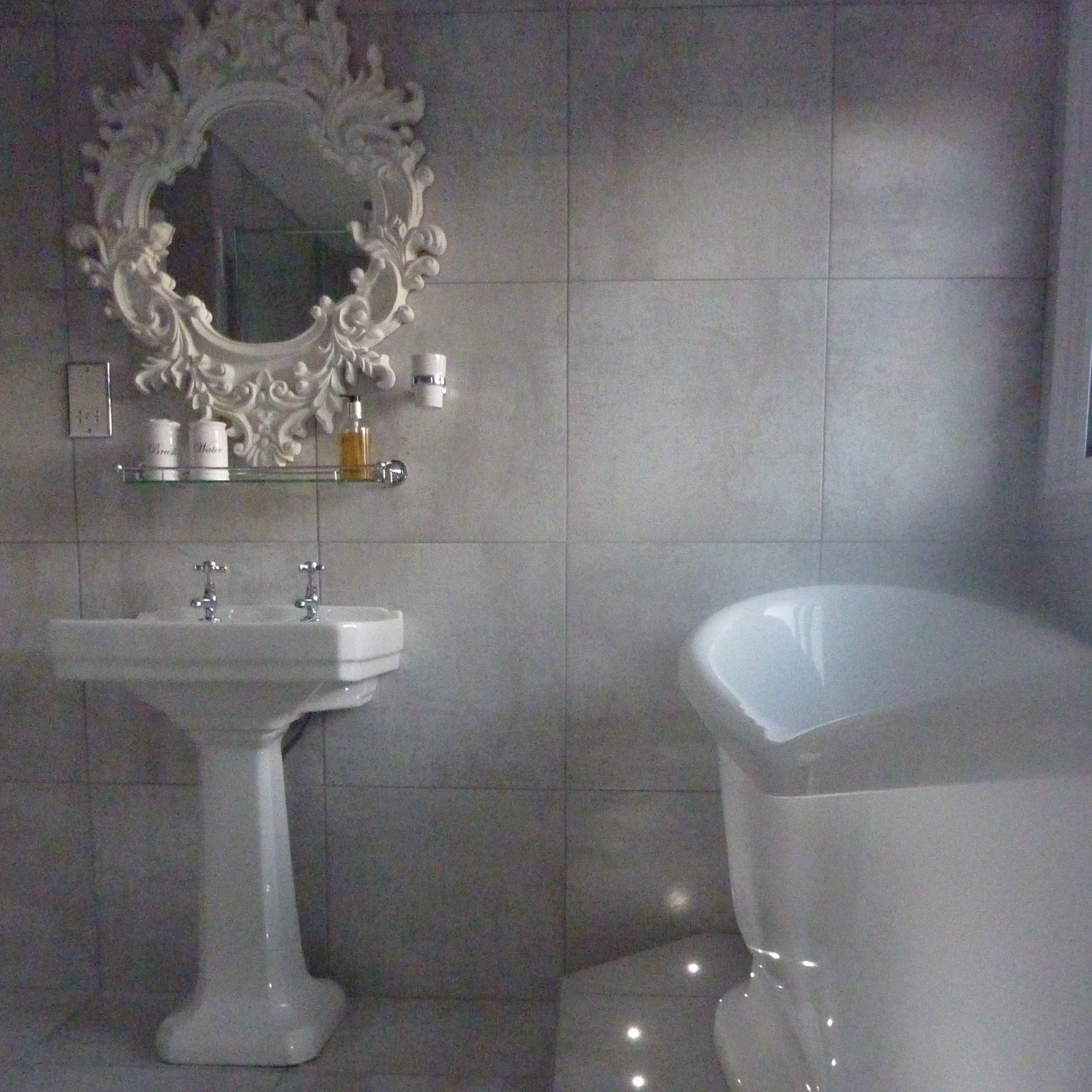 A modern bathroom featuring a freestanding bathtub, a pedestal sink, and a decorative mirror against gray tiled walls.