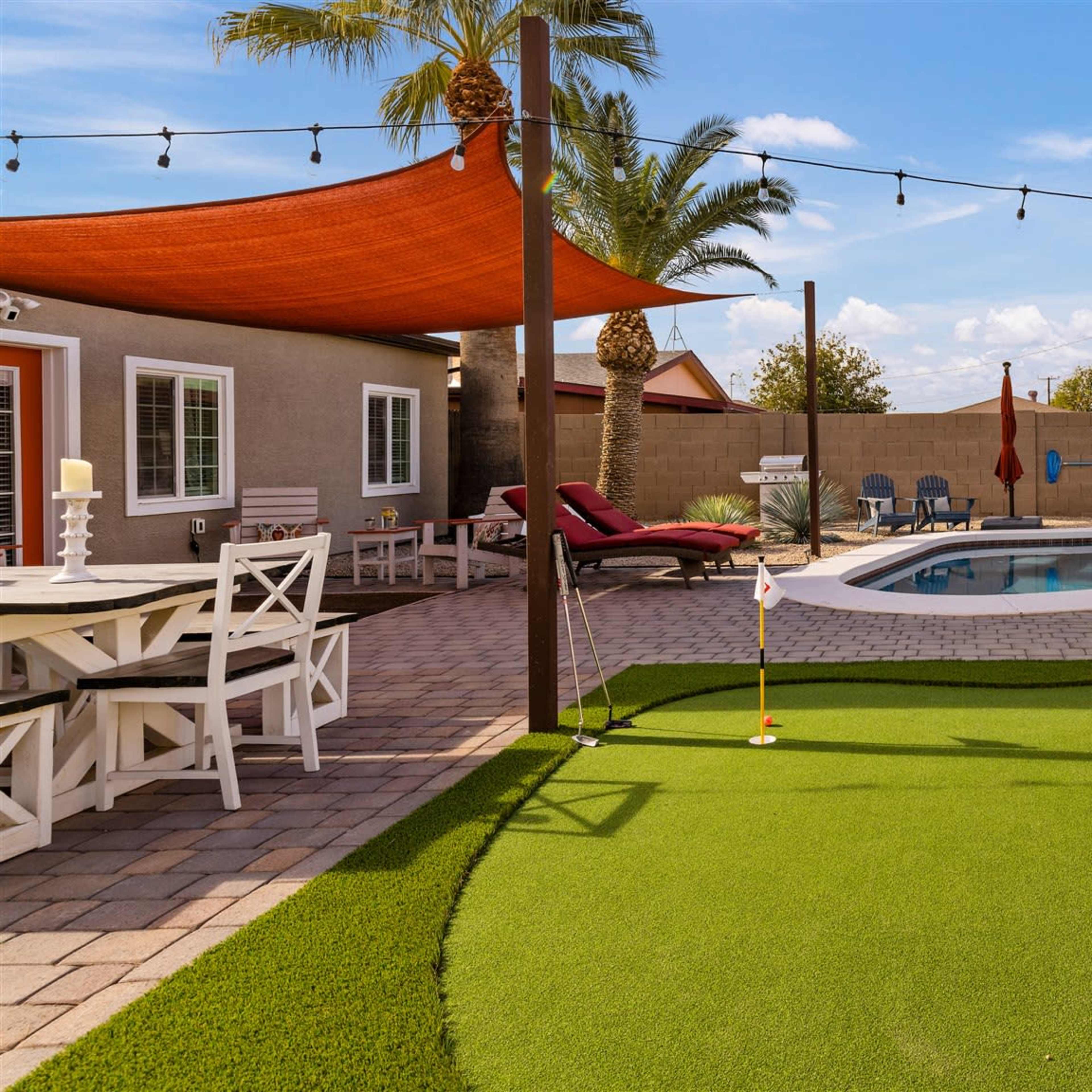 A backyard featuring a putting green, a swimming pool, lounge chairs, and a patio area with a dining table.