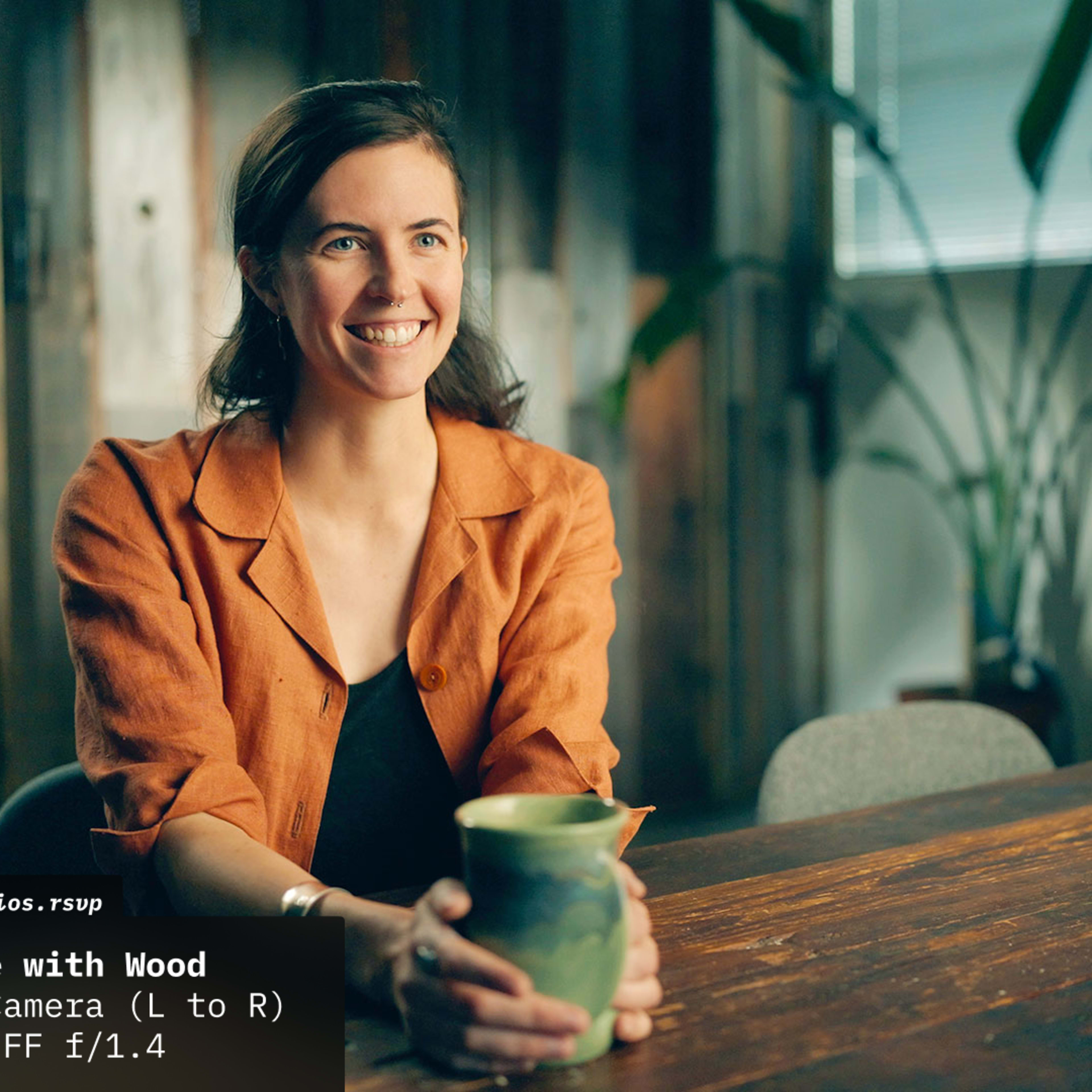 A woman with long hair sits at a table made of wood, holding a ceramic mug, with plants and wooden panels in the background.