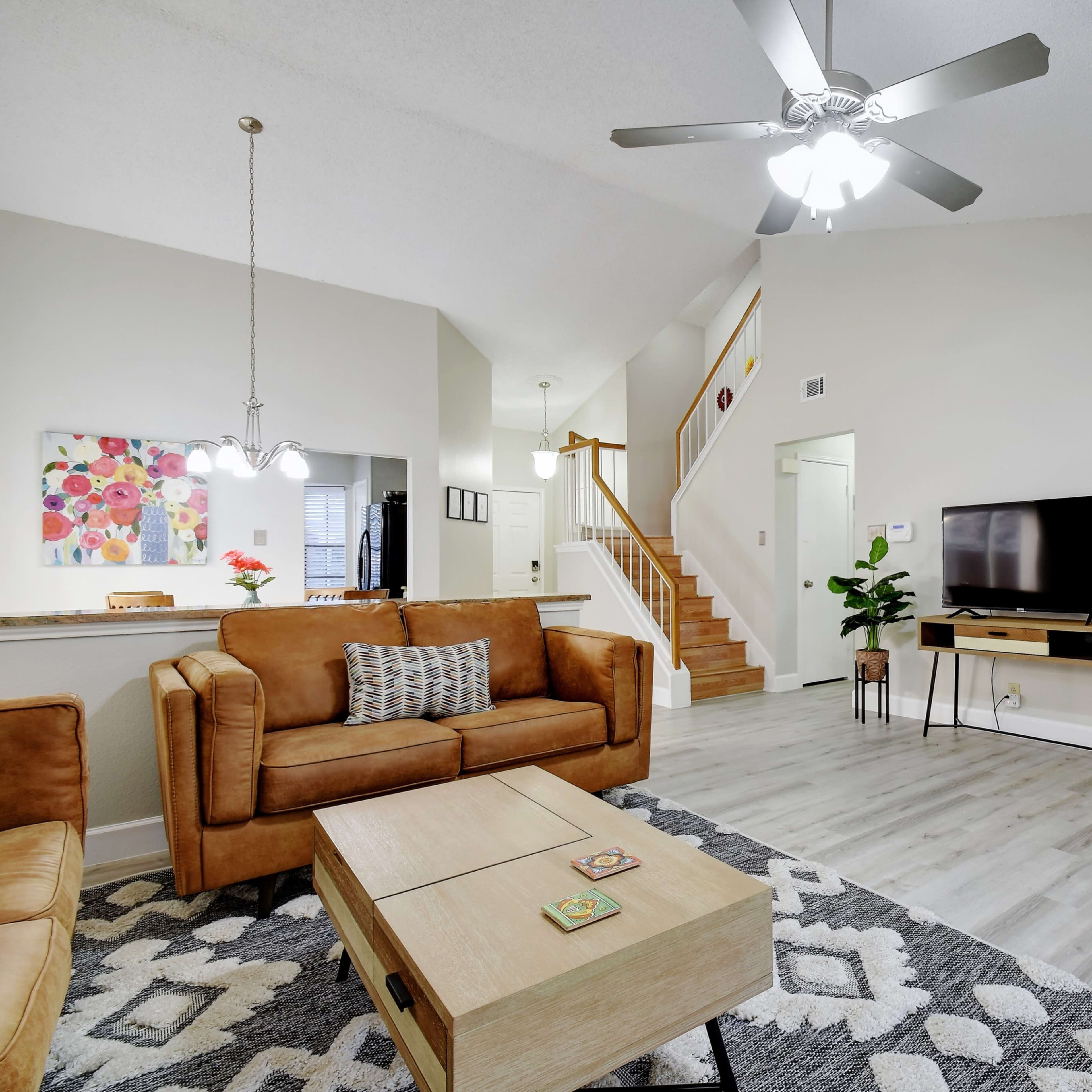 A spacious living room featuring a brown leather sofa, a coffee table, a television on a stand, and a fireplace, with stairs visible in the background.