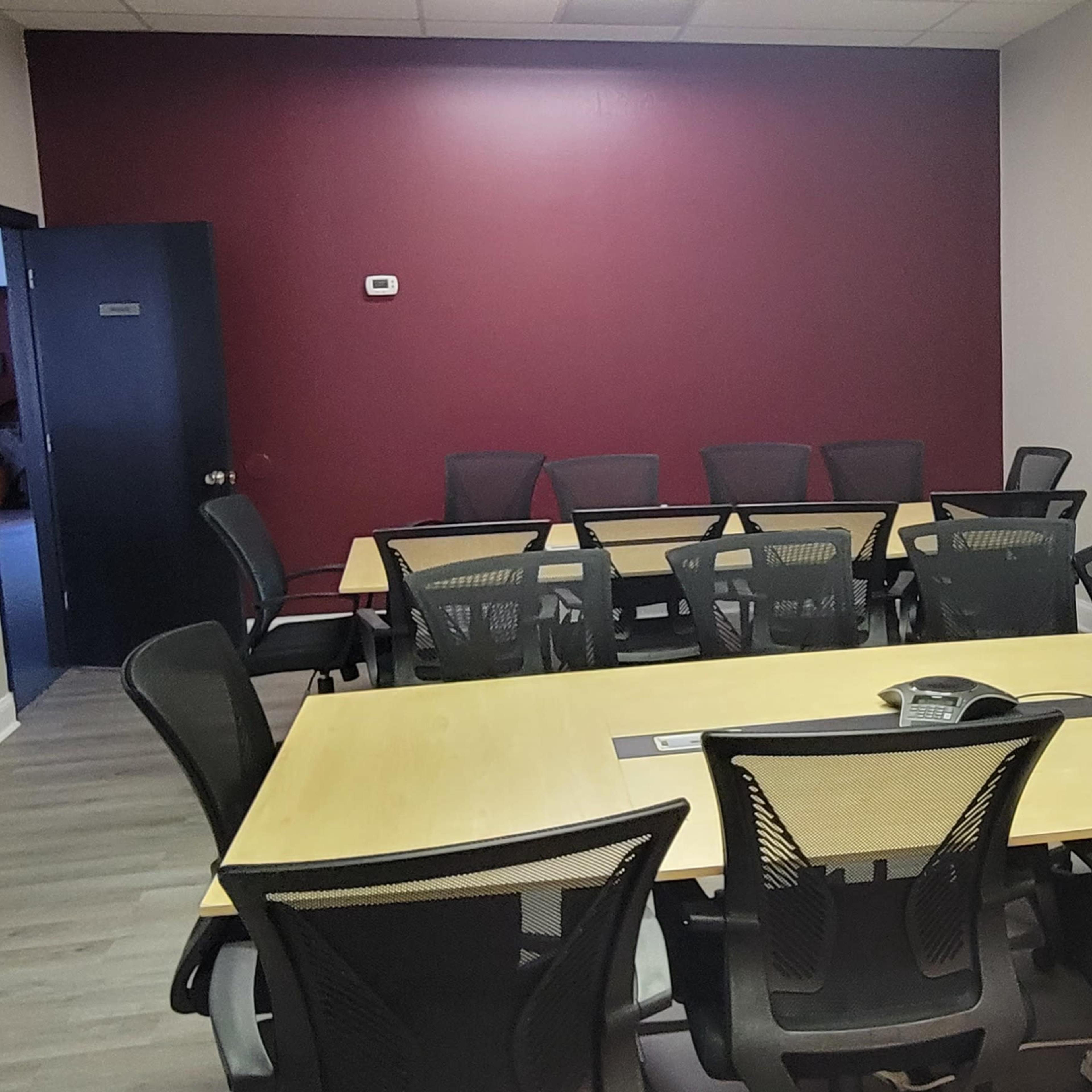 A conference room with a long wooden table surrounded by black mesh chairs, a purple accent wall, and a blue presentation board.