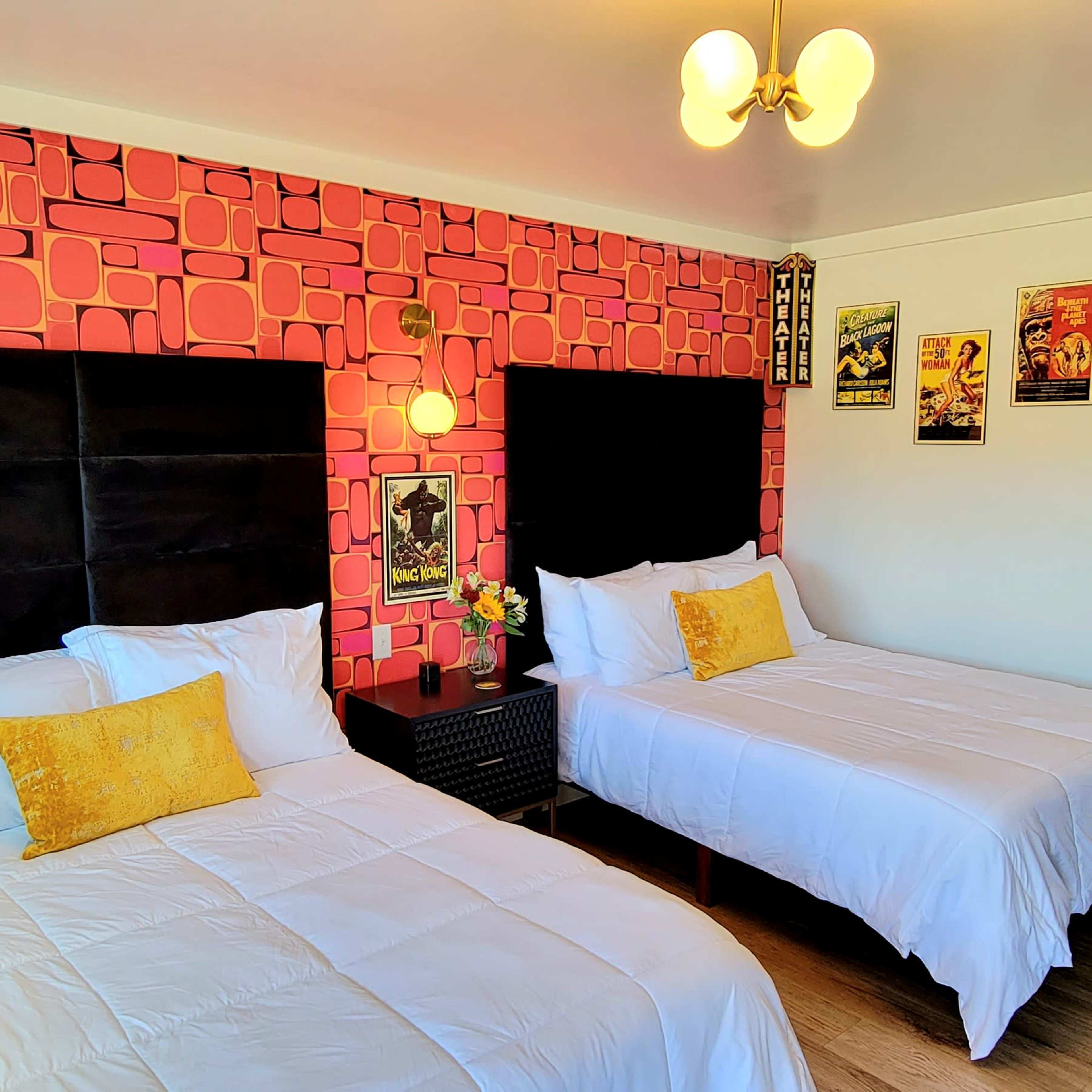 A bright hotel room featuring two beds with white bedding, a patterned pink wall, and decorative wall art.