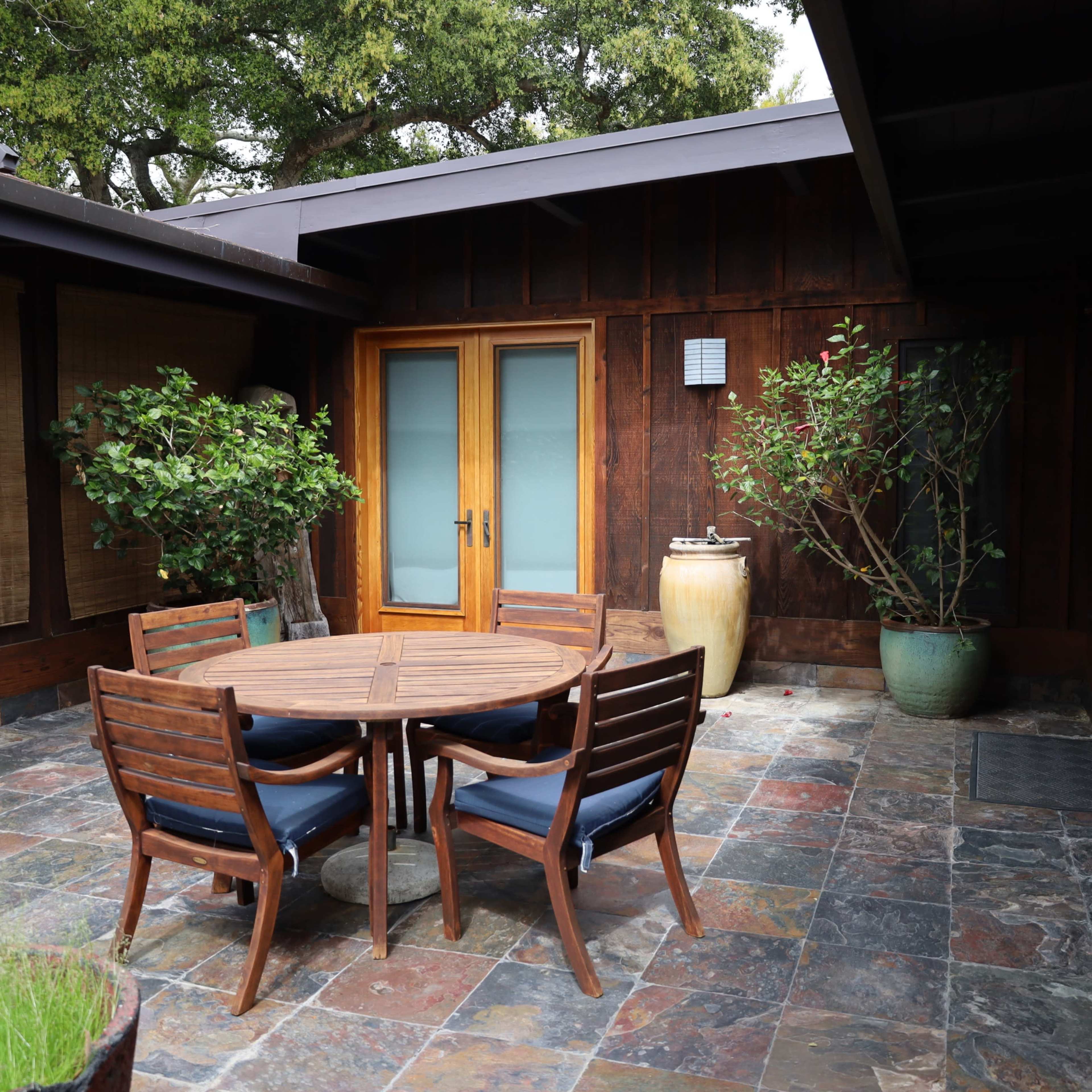 A patio area with a round wooden table surrounded by six chairs, flanked by potted plants and a large ceramic vase.