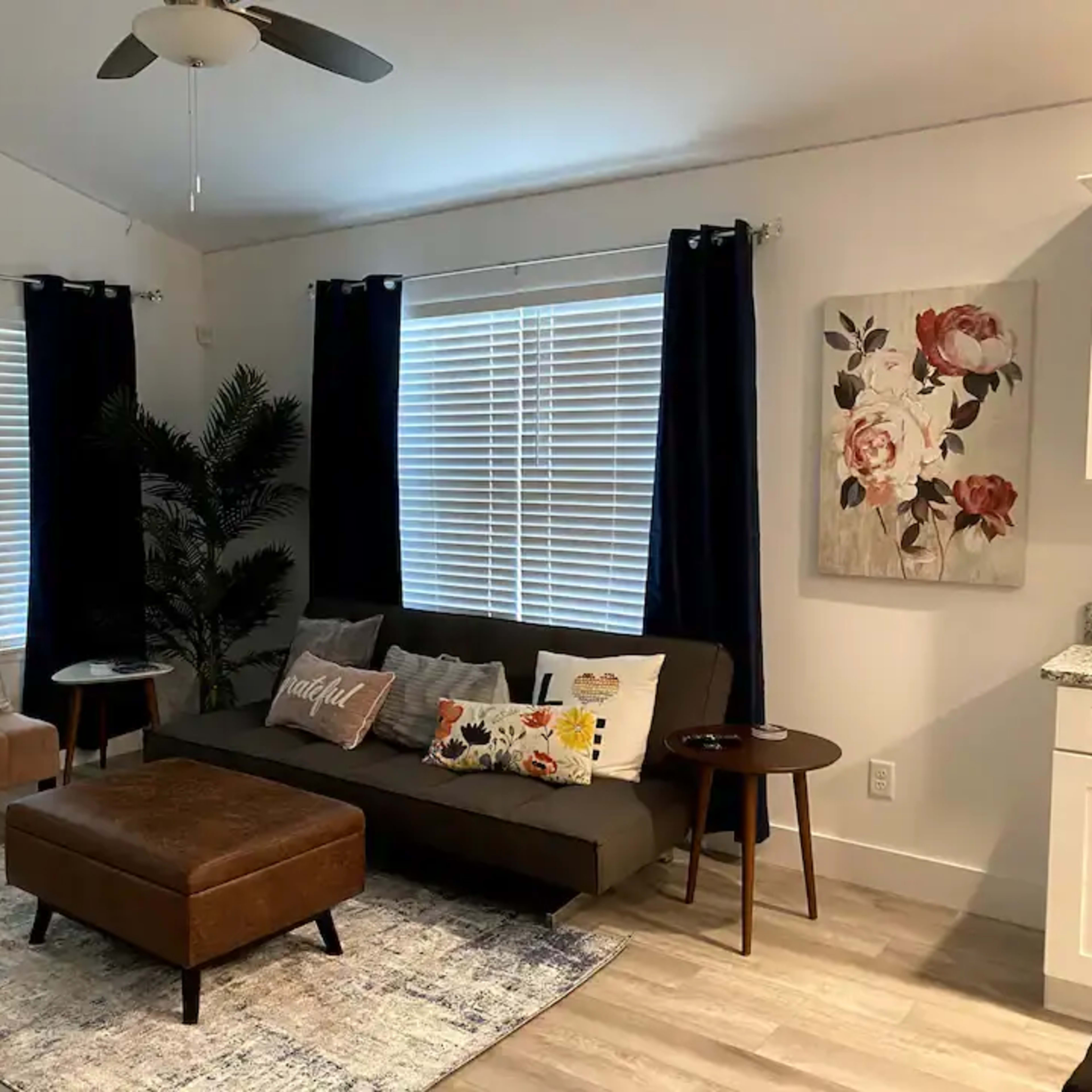 A modern living room with a couch, armchair, and coffee table, featuring large windows with white blinds and dark curtains.