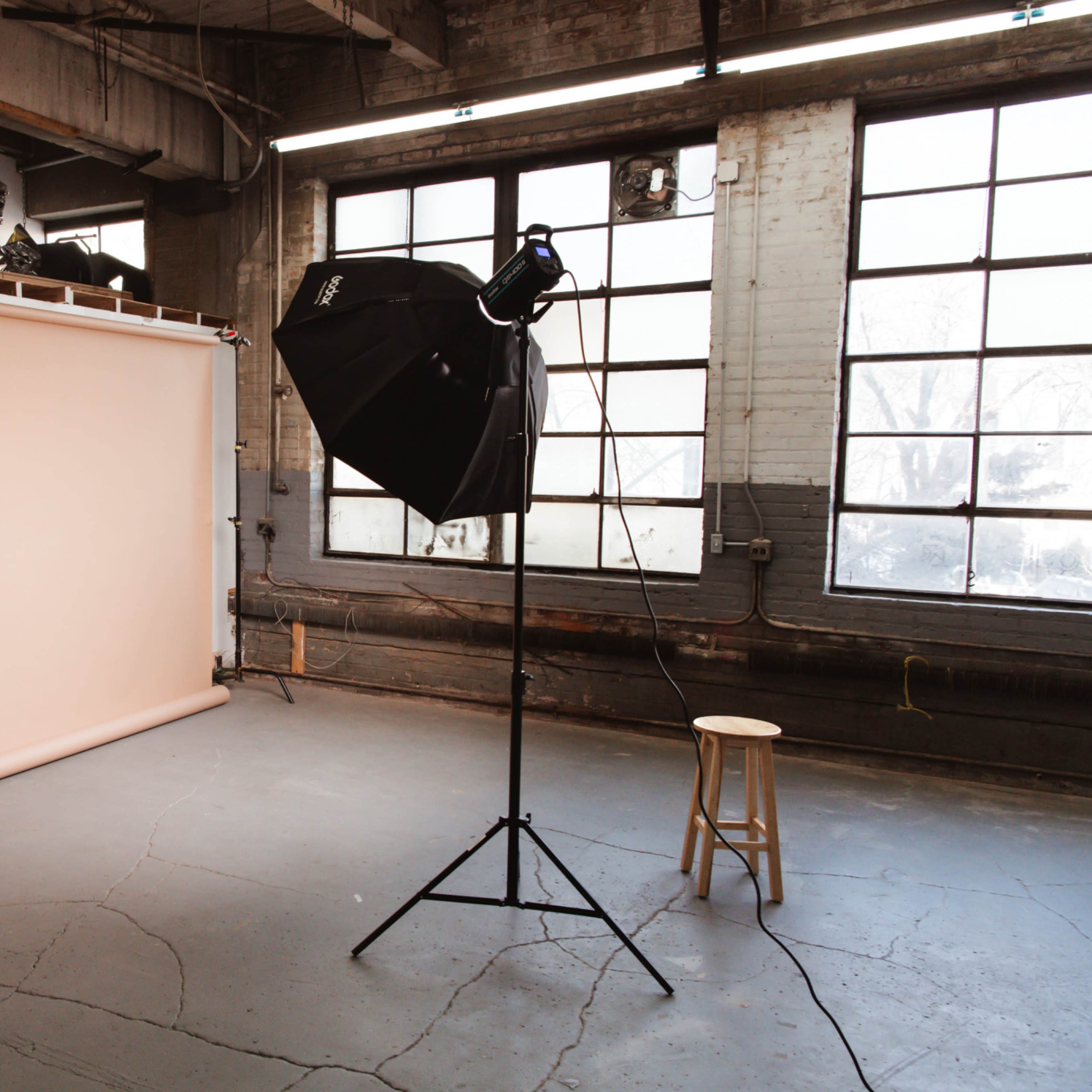 A photography studio featuring a light stand, a beige backdrop, and a stool against large windows.