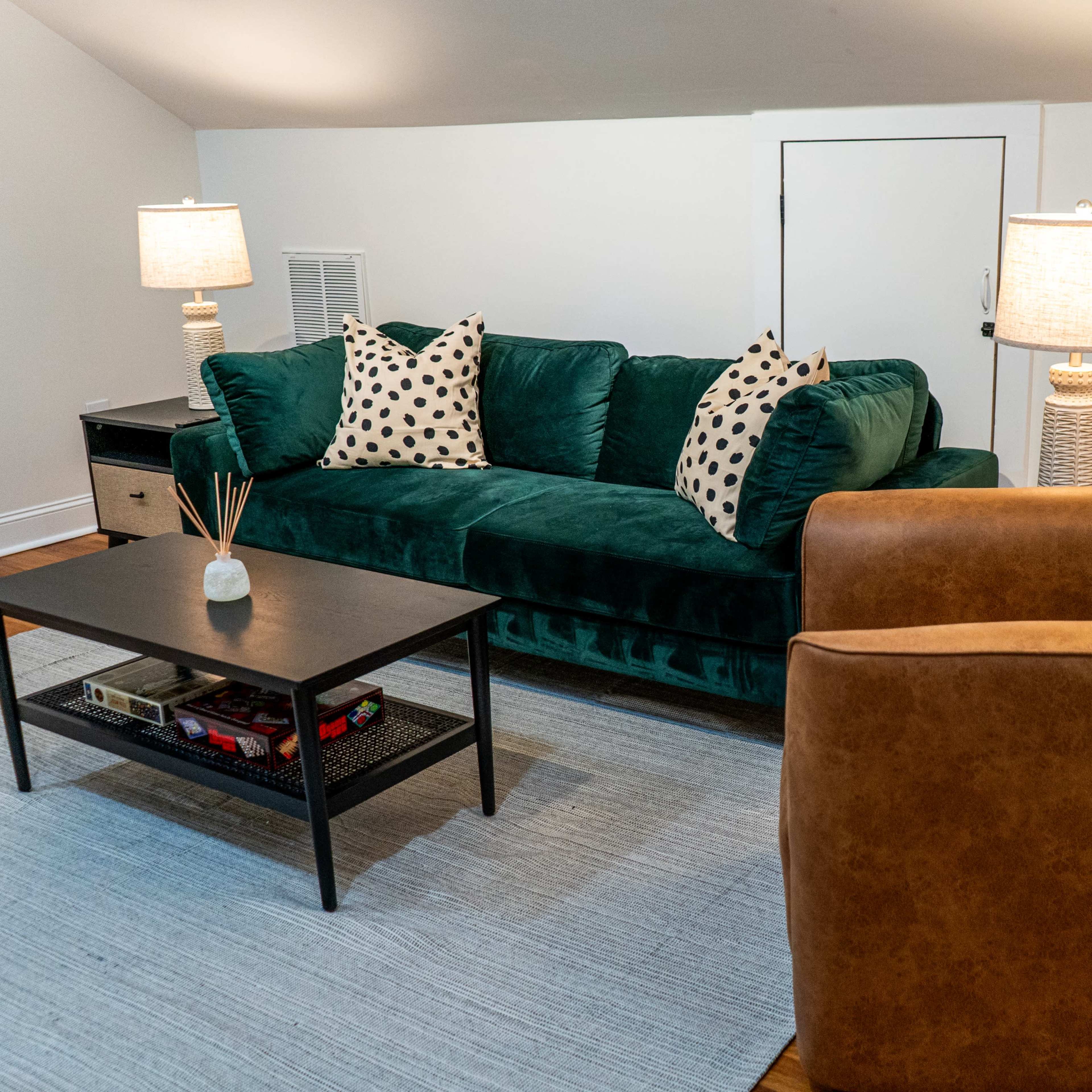 A cozy living room with a green velvet sofa, two lamps, a black coffee table, and a brown armchair, all set on a light-colored rug.