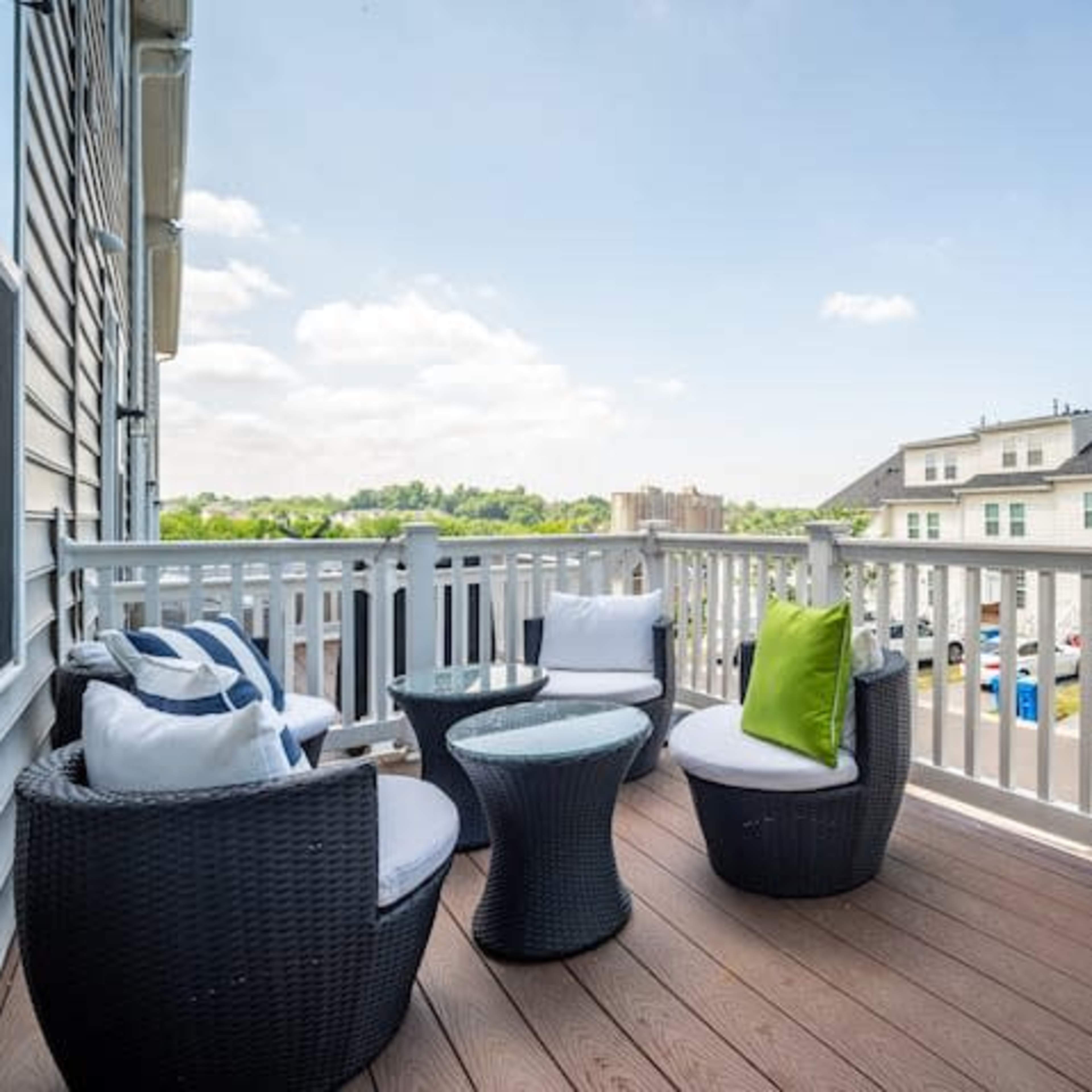 A balcony features four wicker chairs with cushions and a glass table, overlooking a parking area and adjacent buildings.