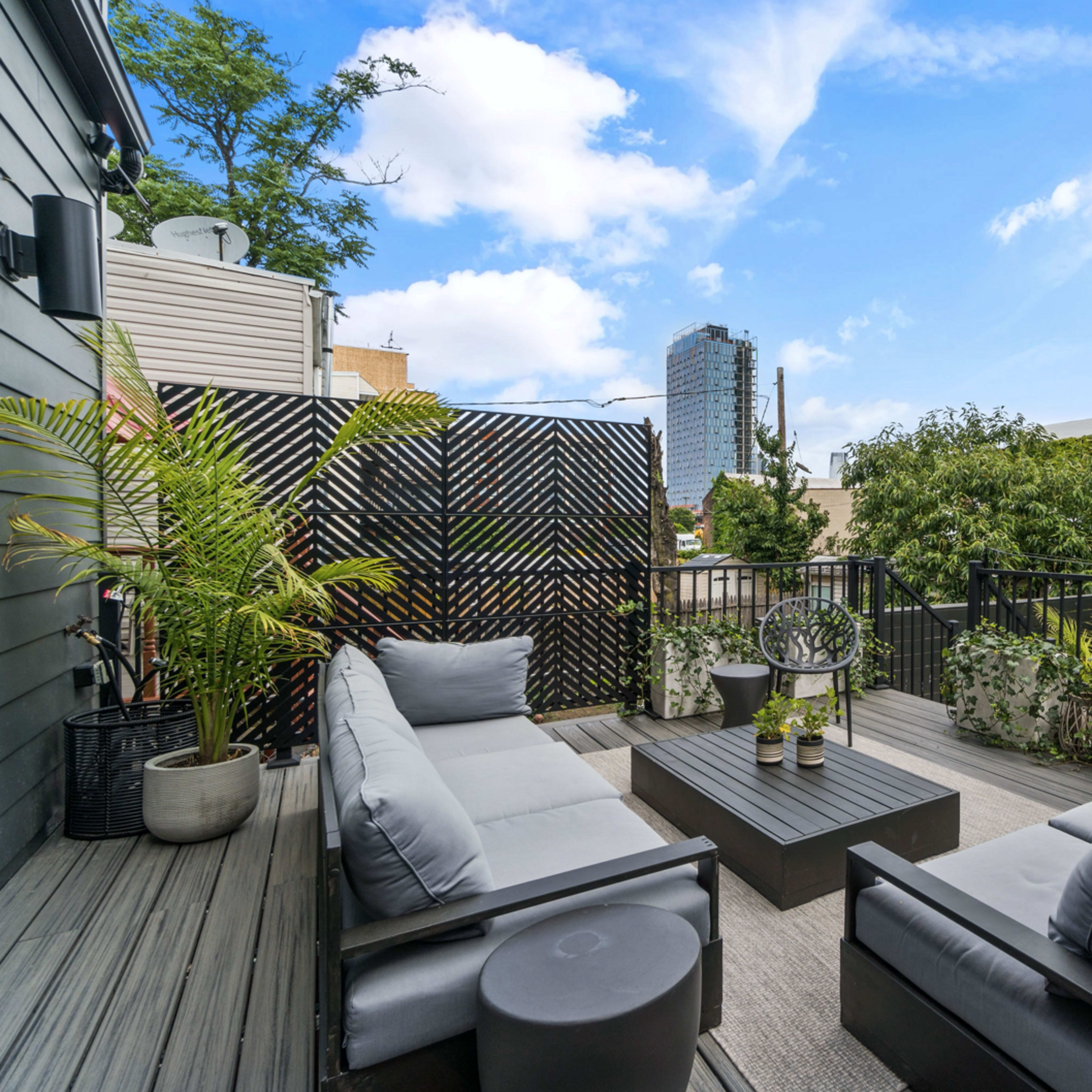 A balcony with gray outdoor seating, a coffee table, and various plants, overlooking a cityscape with tall buildings in the background.